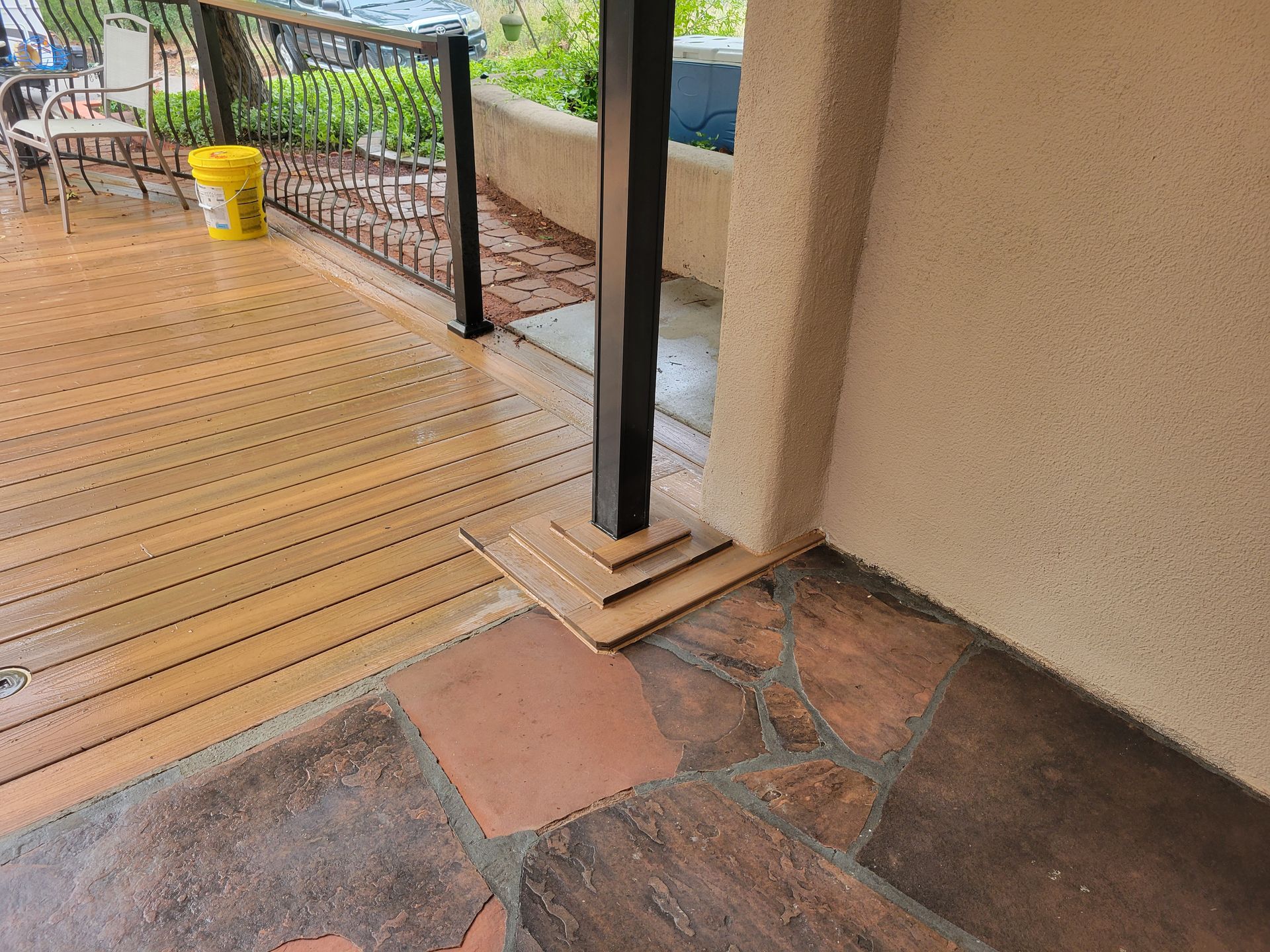 Wooden deck and stone patio with a support post; the post is near a stucco wall and metal railing.