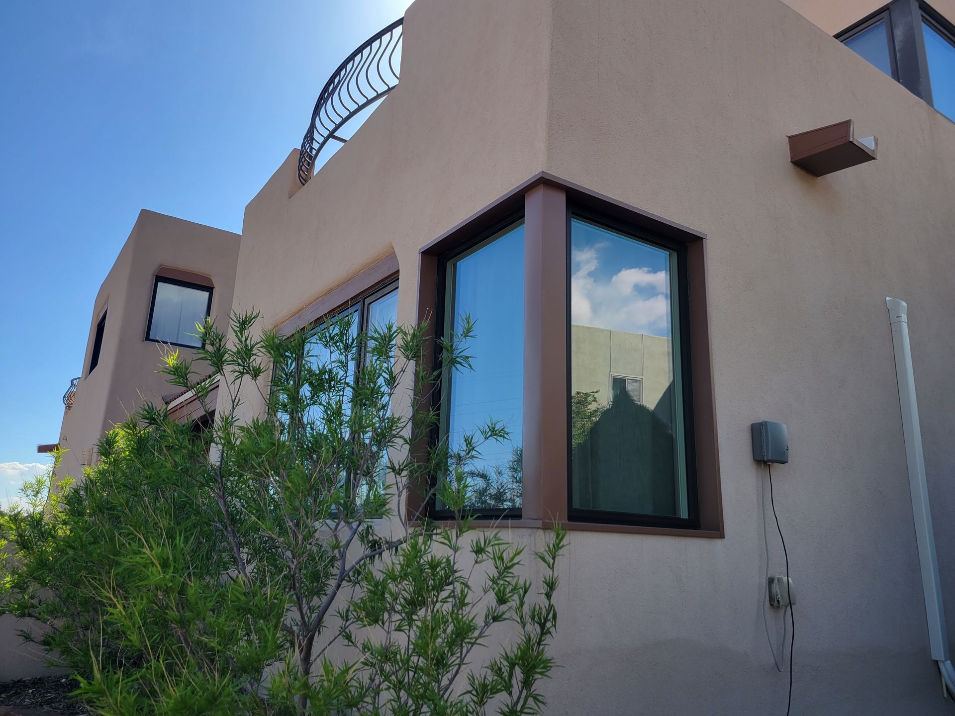 Beige stucco building with dark-framed windows, bronze trim, and a wrought iron balcony. Green shrubbery in foreground.