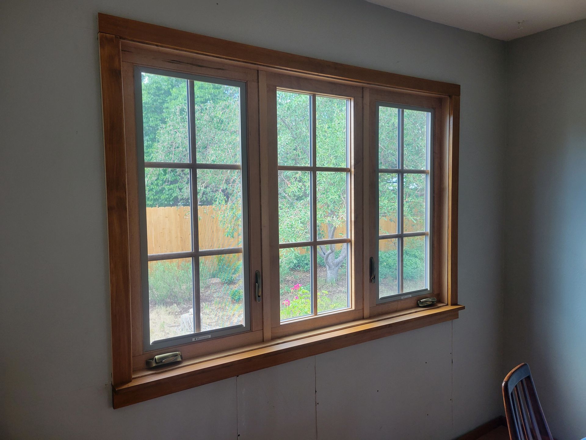 Wooden-framed window with three panes, looking out onto a backyard. Light brown trim and wall.