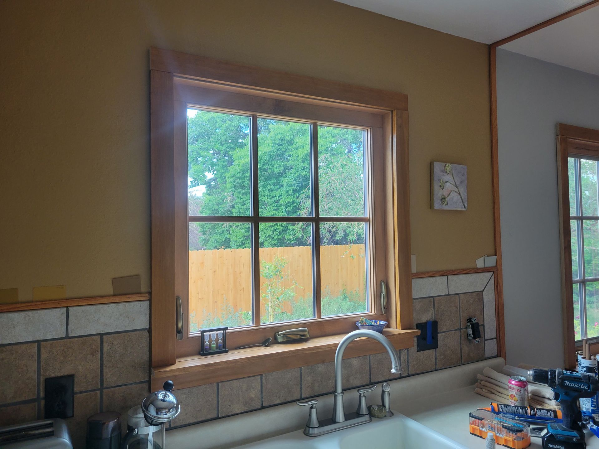 Kitchen window with wood trim, sink, and tiled backsplash; view of trees outside.