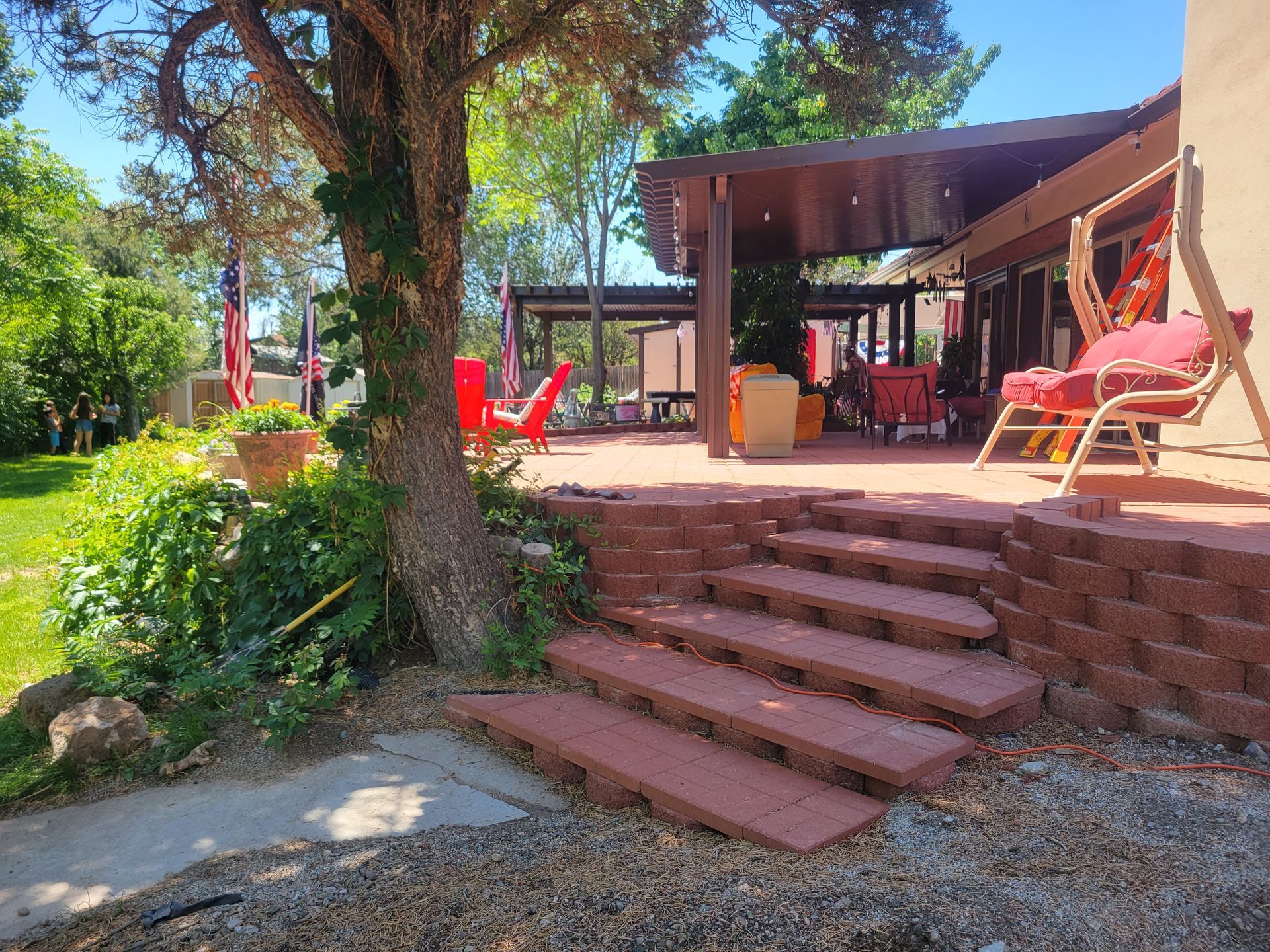 Brick steps lead to a patio with red chairs and a covered area, next to a tree, under a blue sky.