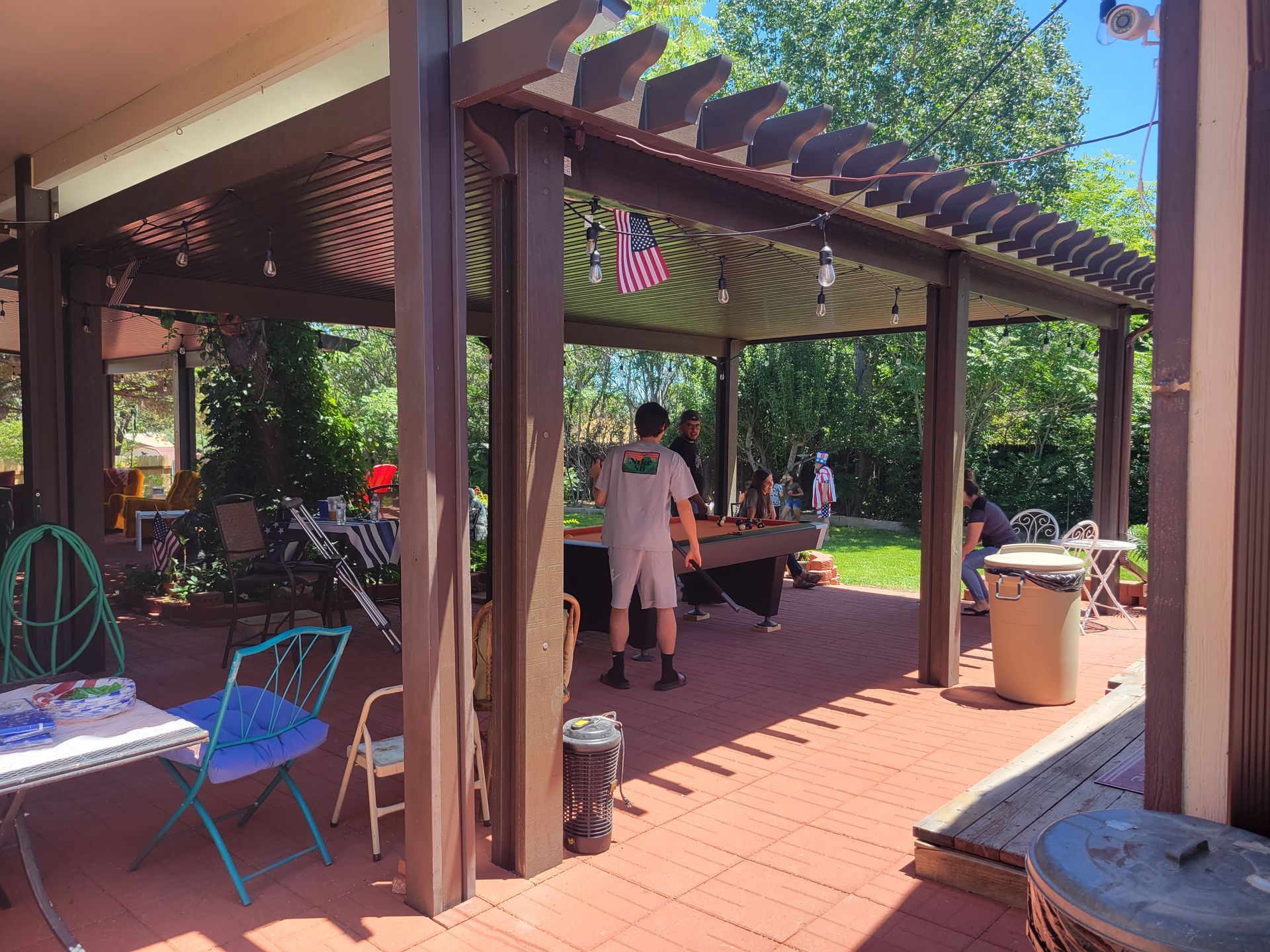 Outdoor patio with pergola, people gathered around a table; brick flooring, green lawn, sunny day.