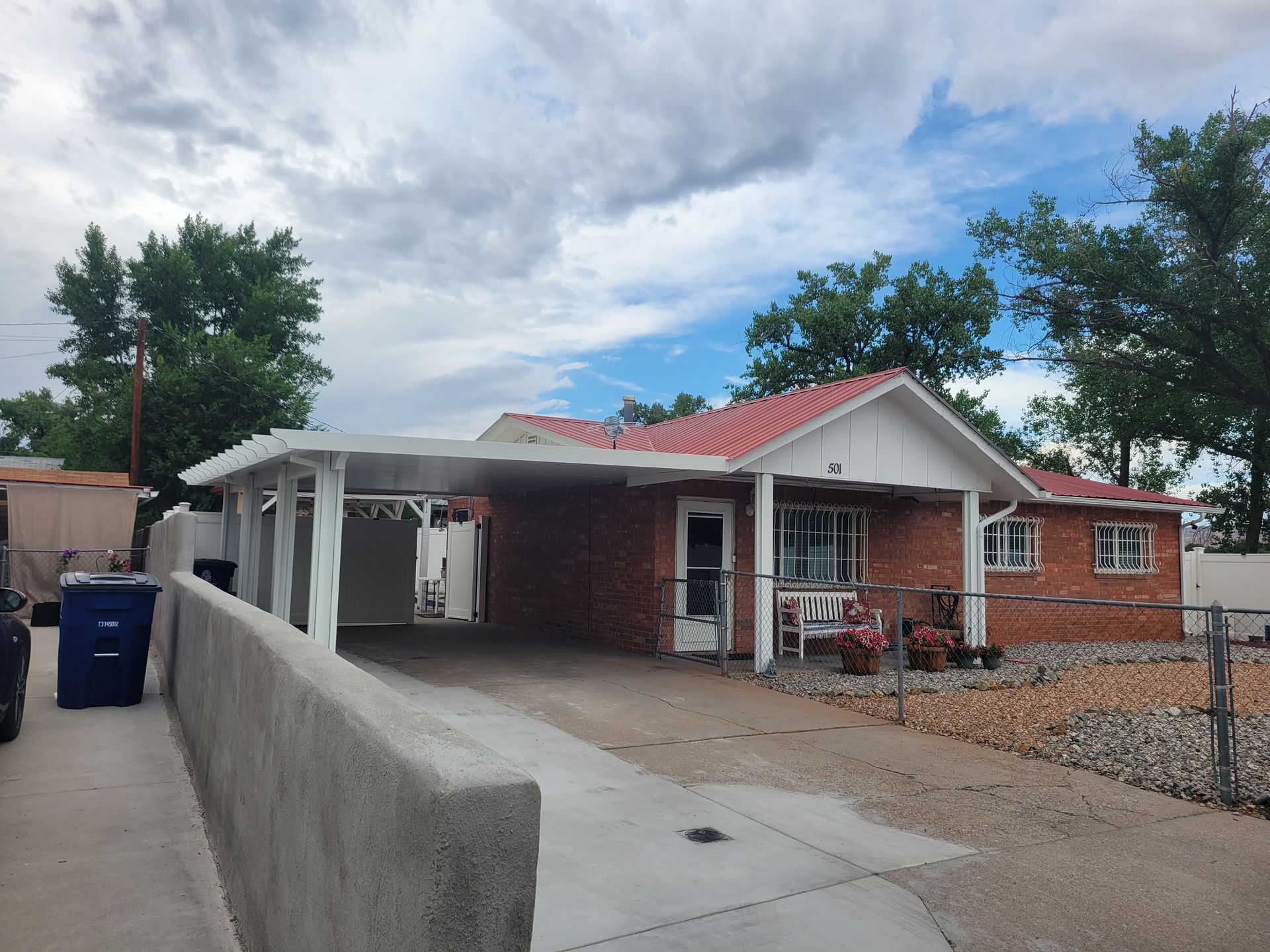 Brick house with a red roof and carport, concrete driveway, fence, and cloudy sky.