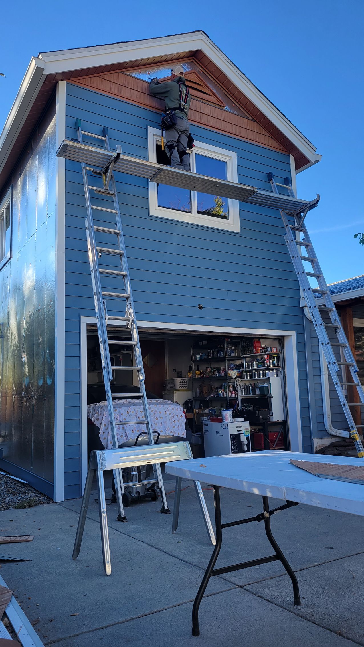 Two workers on ladders repairing blue-sided building with open garage; clear sky.