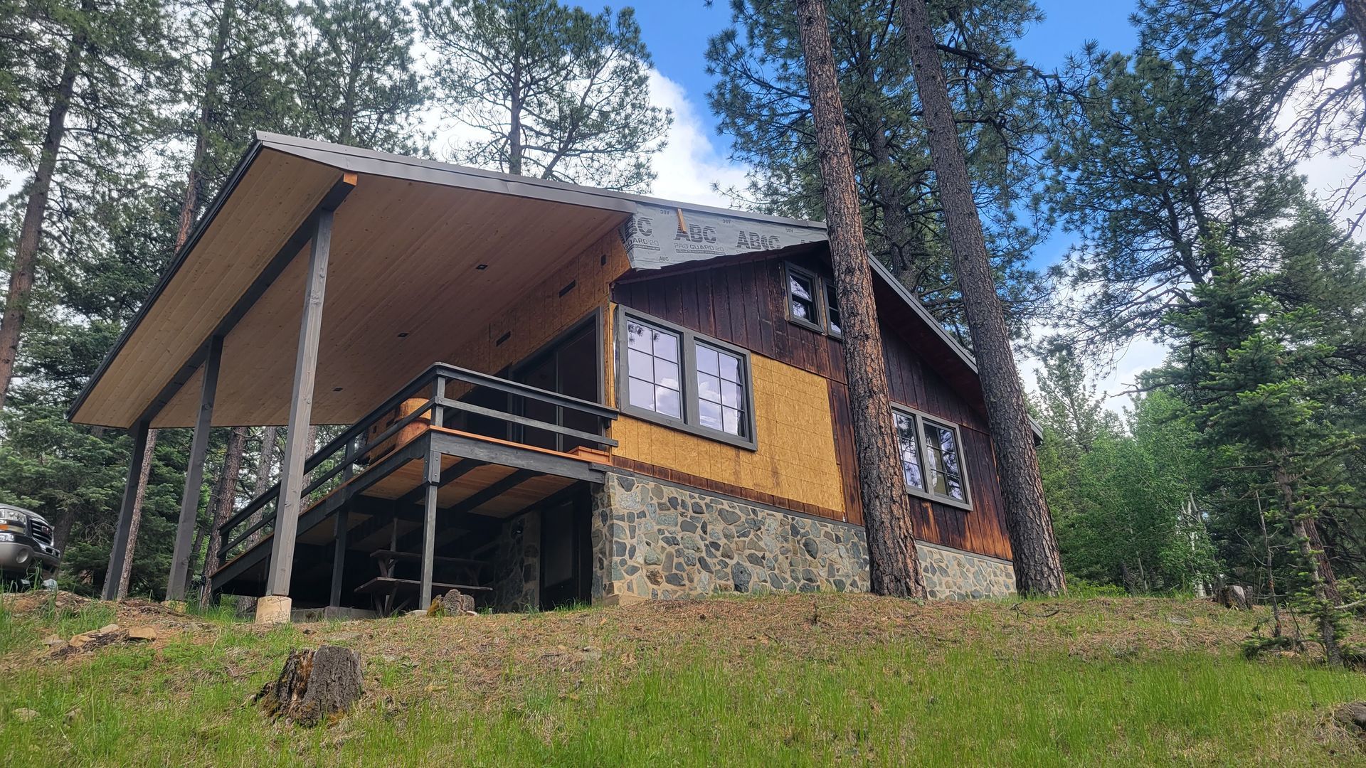Cabin with porch, stone base, in a wooded area with tall trees and green grass.