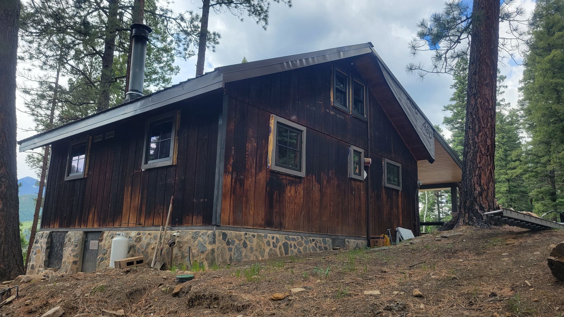 Wooden cabin in a forest setting. Brown exterior, stone foundation, metal roof. Windows, chimney visible.
