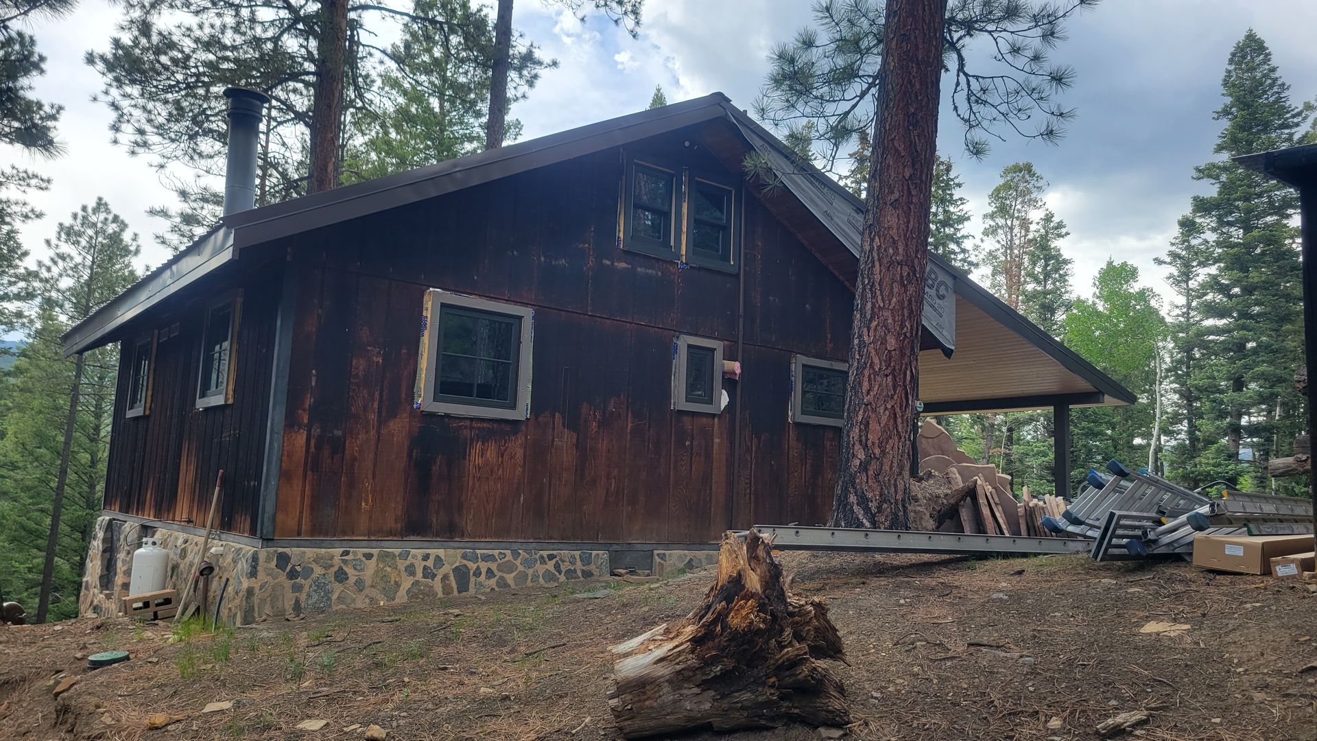 Rustic cabin in a forest setting. Brown exterior, stone foundation, trees in background, overcast sky.