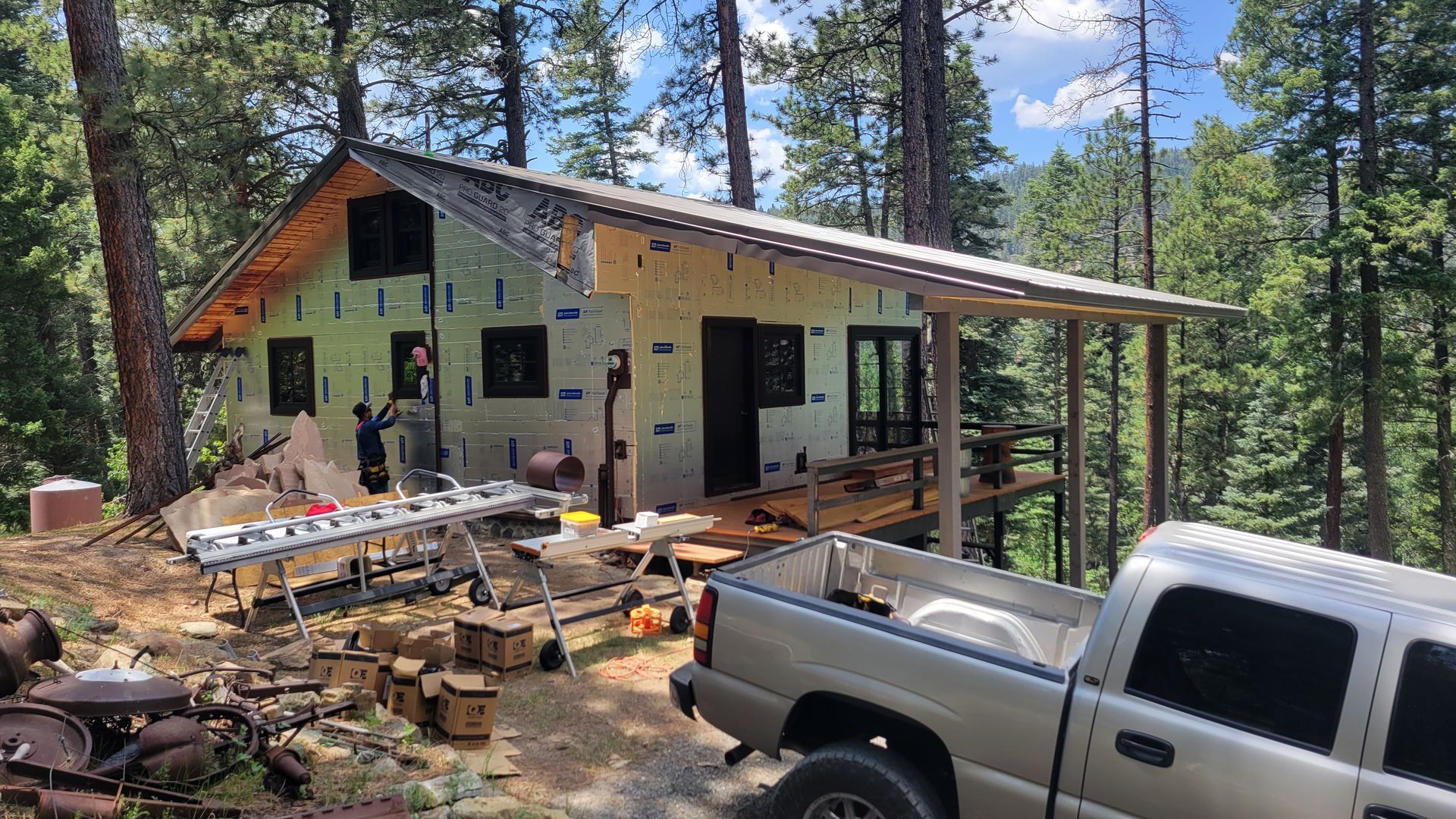 A house under construction in a forest setting. A pickup truck is parked nearby.