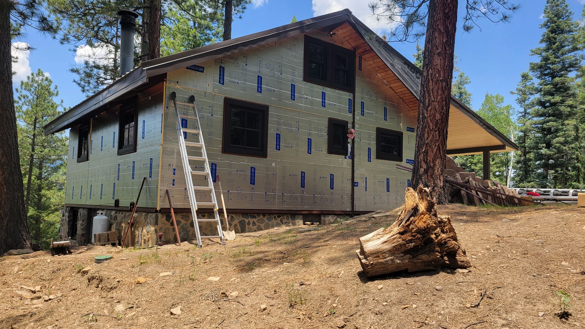 Cabin under construction in a wooded area, with insulation visible. A ladder leans against the structure.