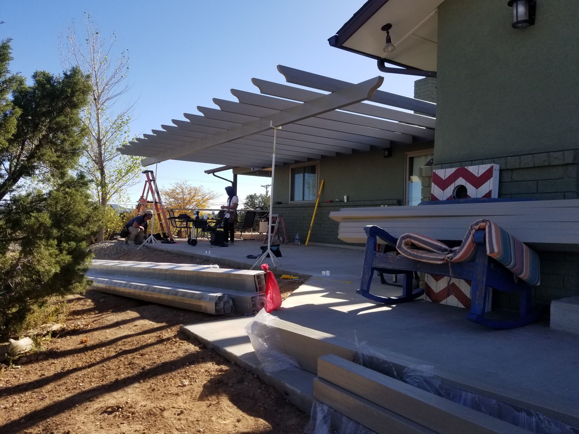 Pergola extending from a green house with stacked building materials and a rocking chair on the porch.