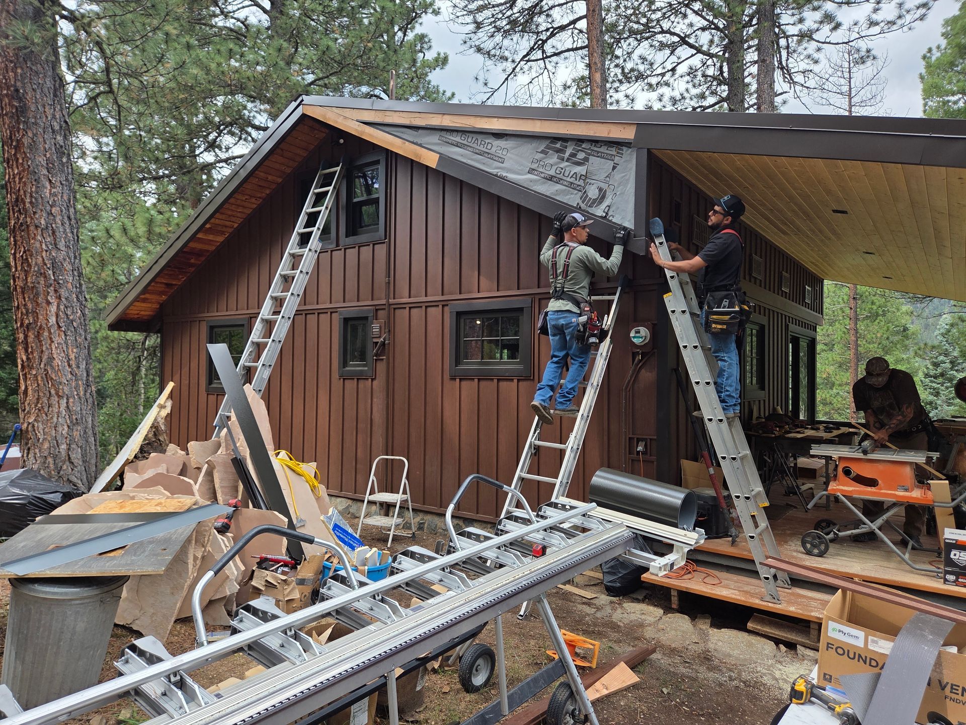 Construction workers on ladders installing siding and roofing on a brown cabin in a wooded area.