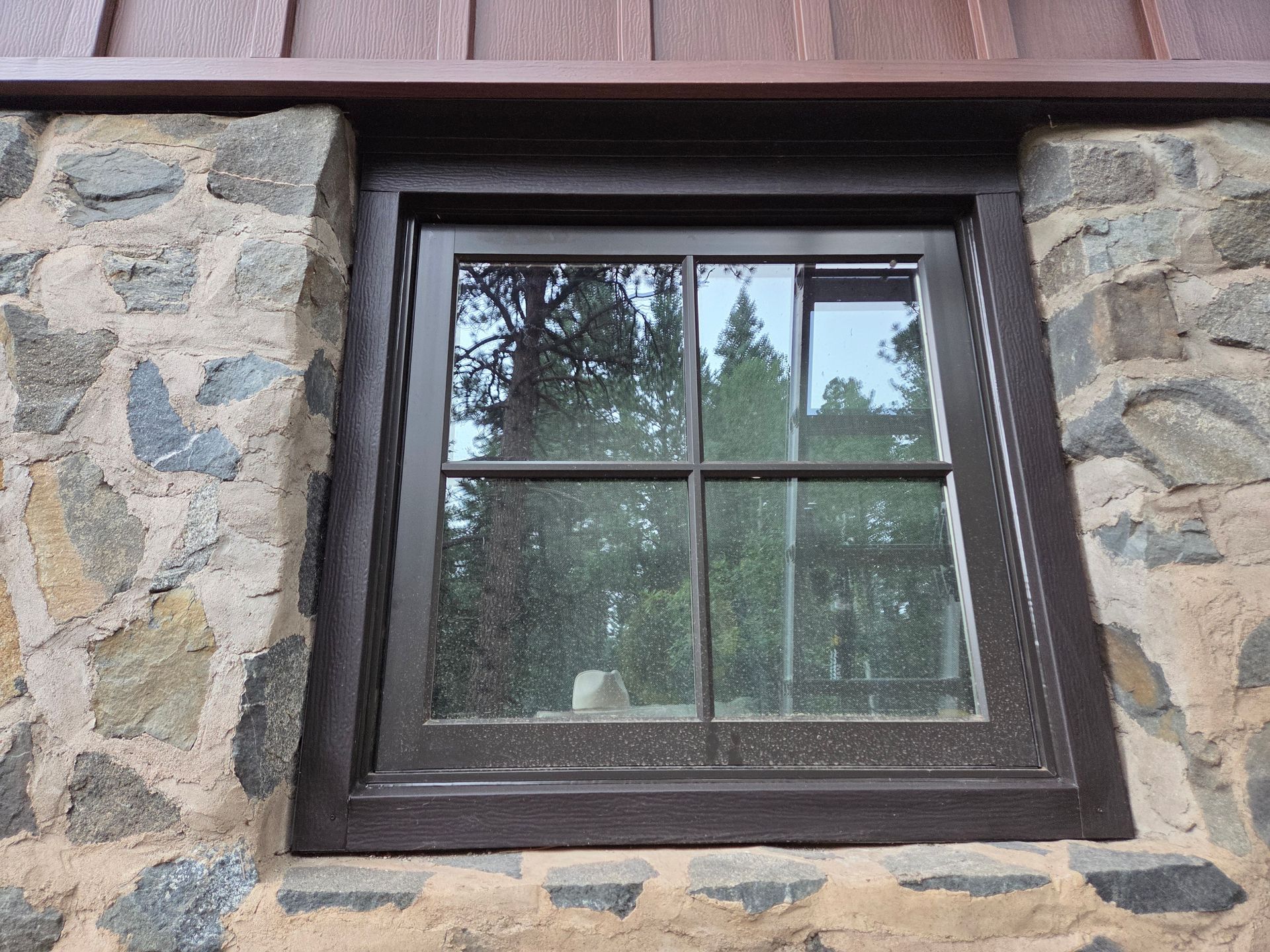 Brown-framed window set in a stone wall. Trees are reflected in the glass.