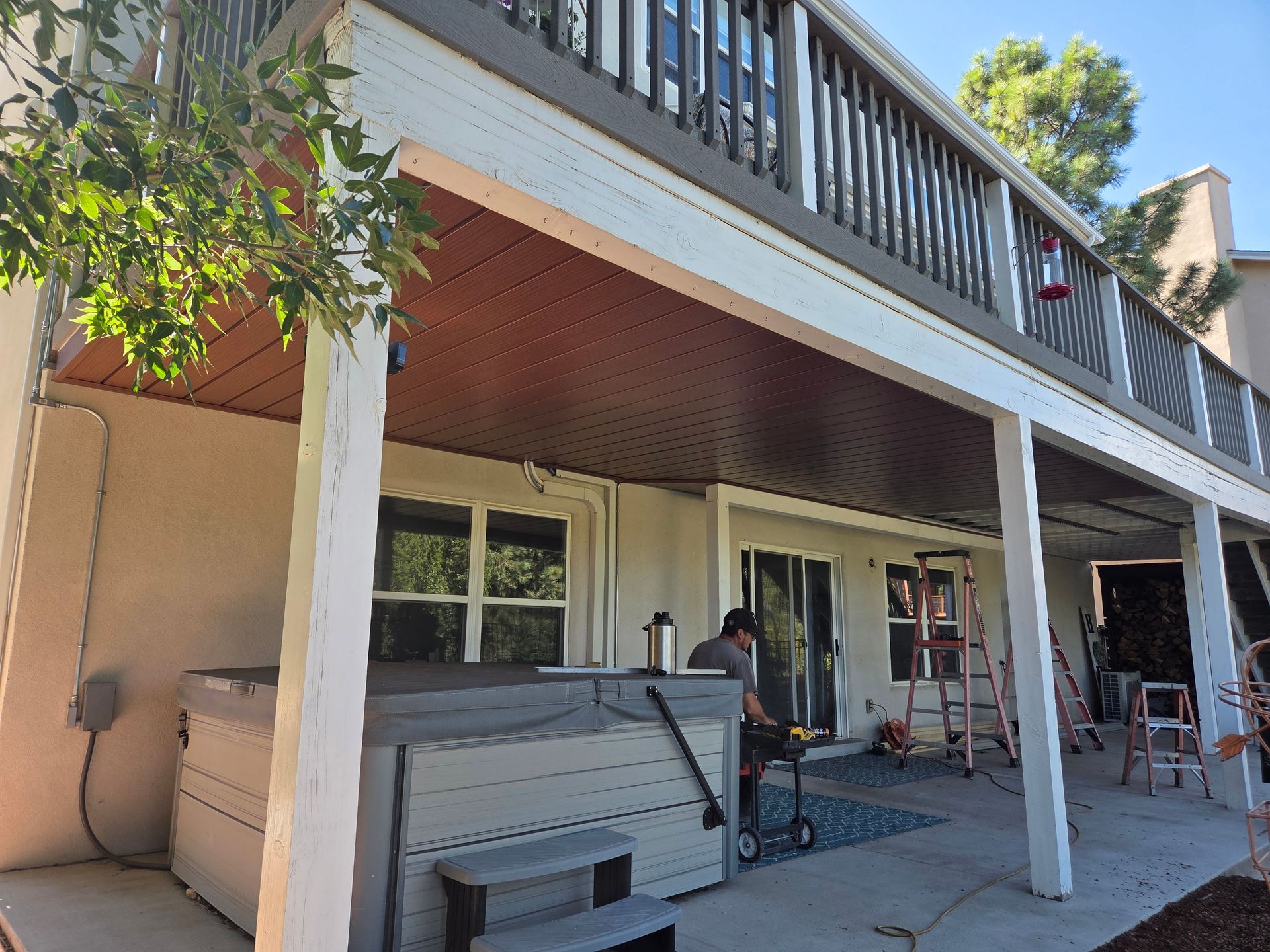 Two-story house with a deck. A person works on the deck below. A hot tub is visible.