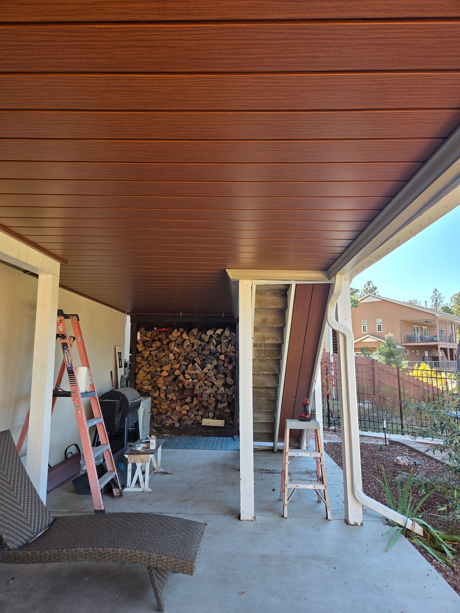 Covered patio with wood-look ceiling; firewood stacked behind supports; ladders present.