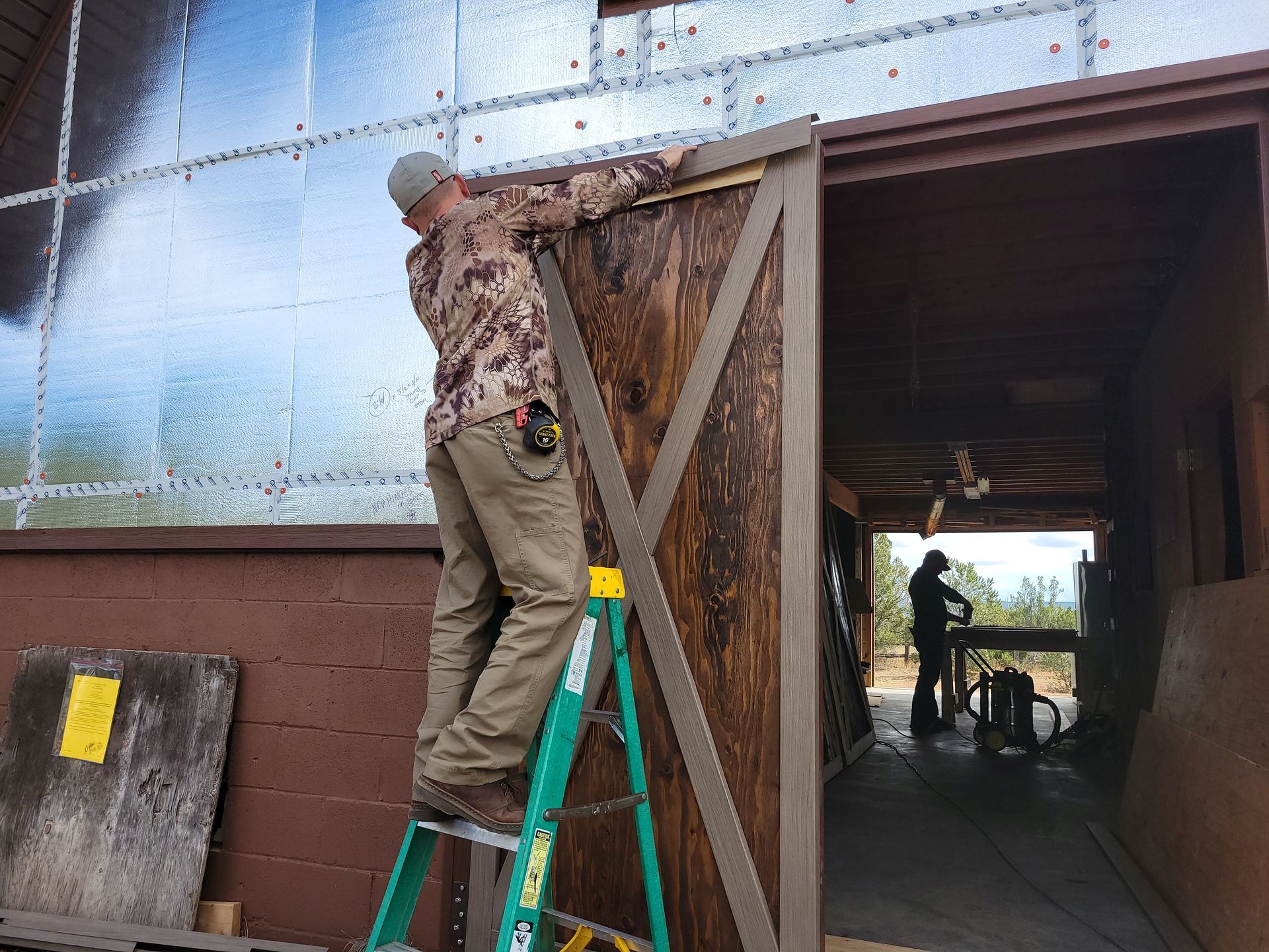 Man on a ladder installing a wooden barn door on a brown building with metallic siding.