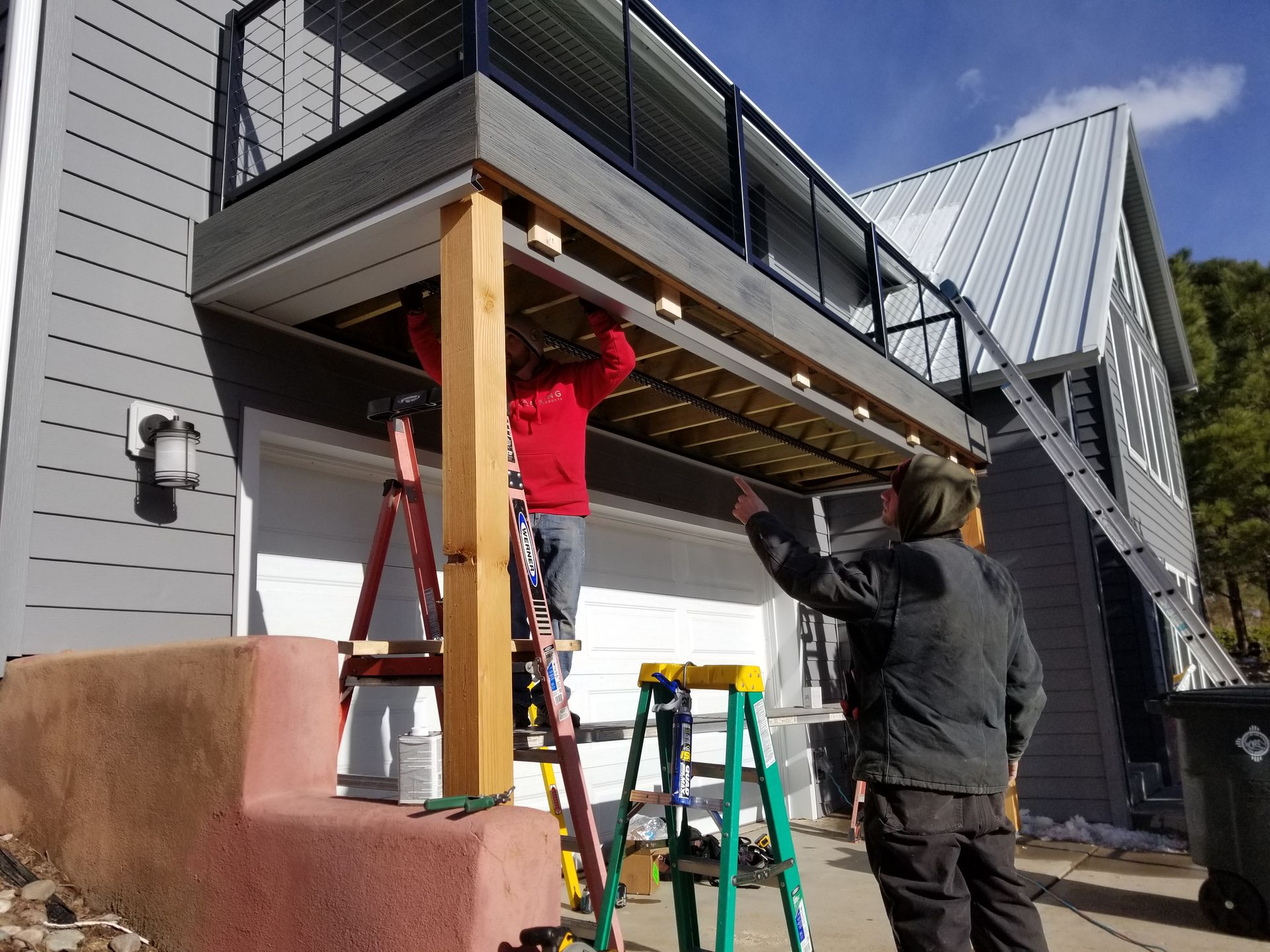 Two people working on deck construction near a gray house with a metal roof. One on a ladder, another pointing.