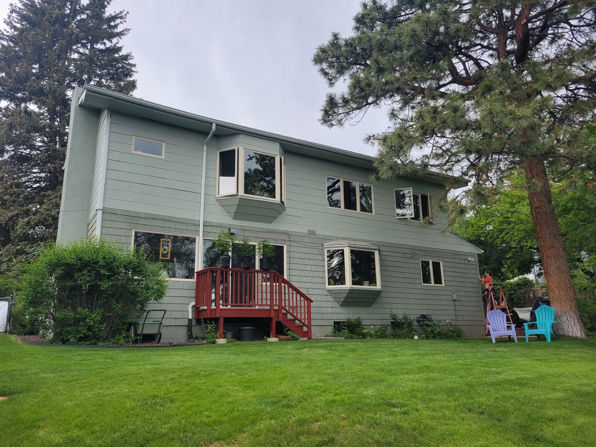 Green two-story house with red deck, large tree, and chairs on a grassy lawn. Overcast sky.