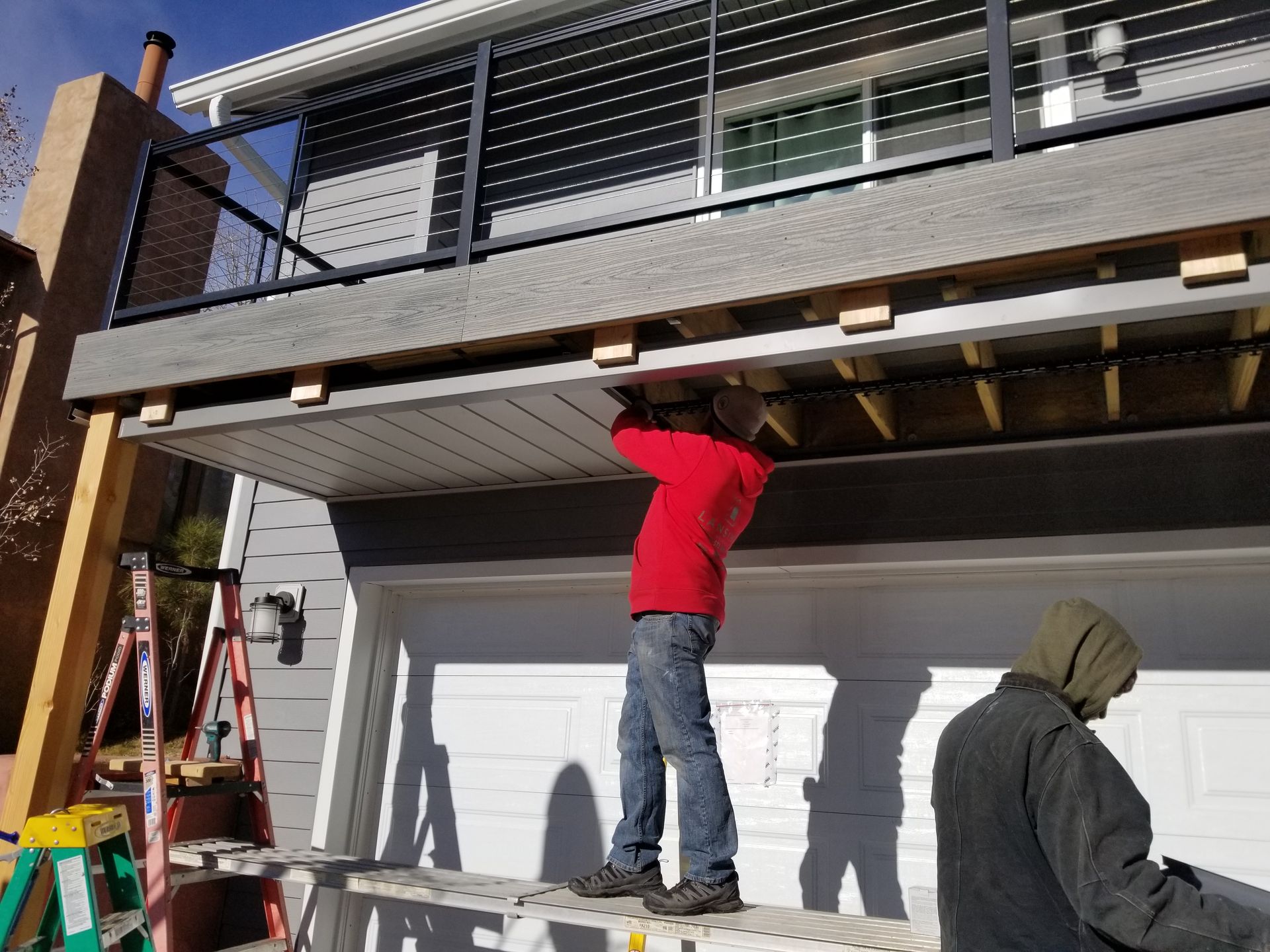 Construction workers installing siding under a deck above a garage. One on a ladder, another observing.