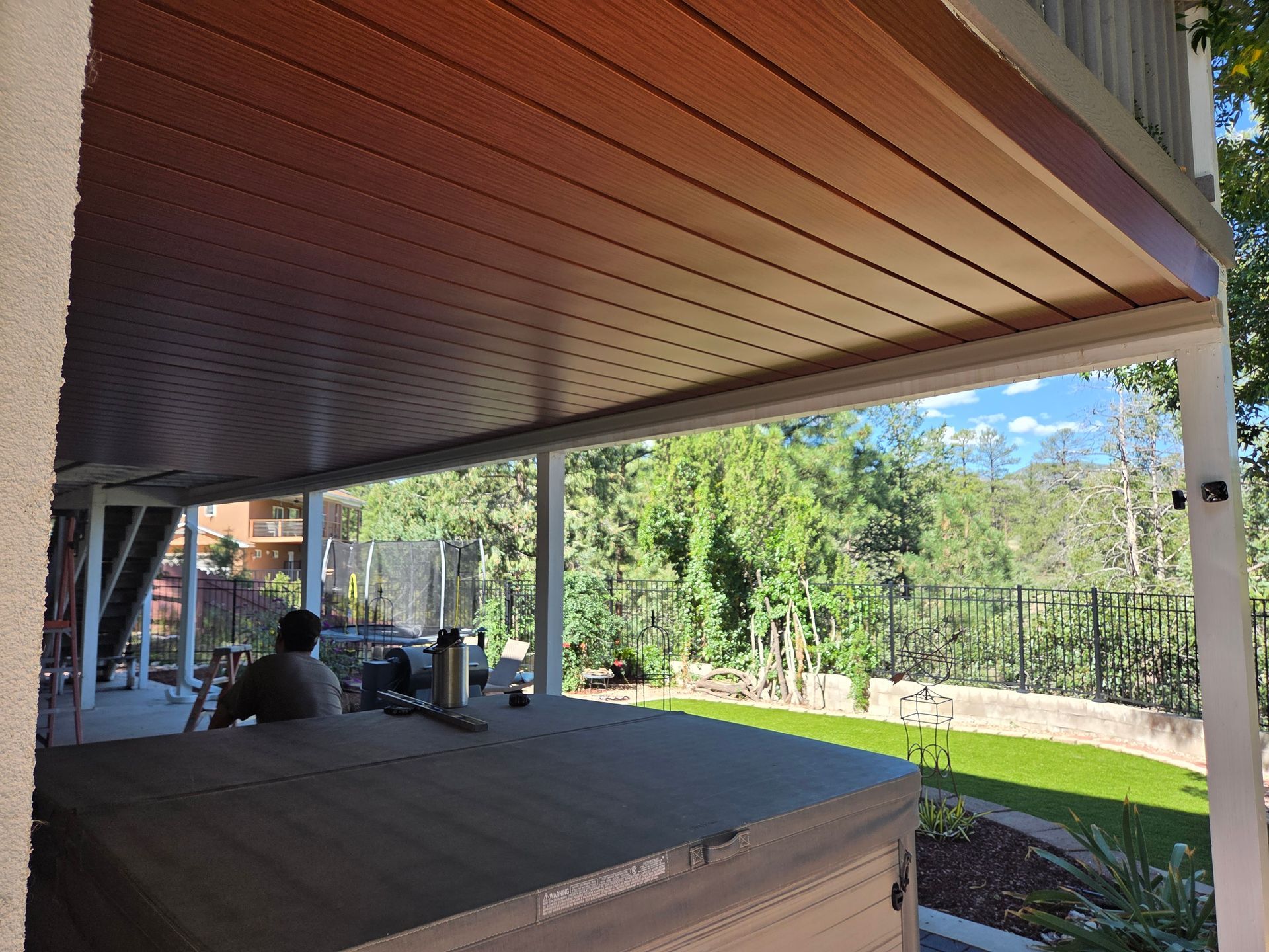 Covered patio with a brown ceiling, overlooking a backyard with a hot tub and trees.