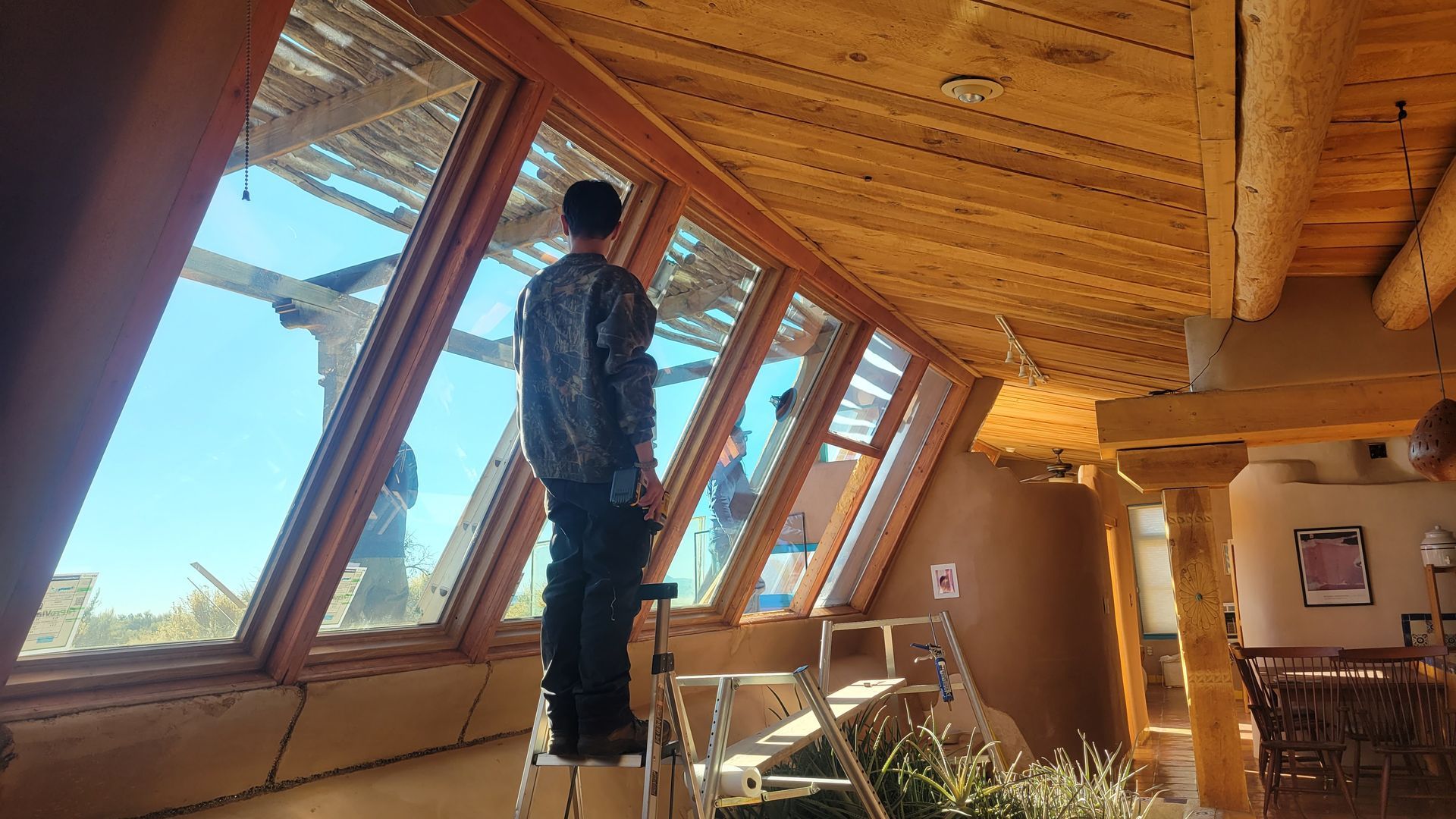 Man on a ladder looks out slanted windows of a wood-beamed building. Sunlight streams in.