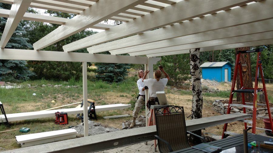 Two people building a white pergola outdoors. Green trees and a blue shed are in the background.