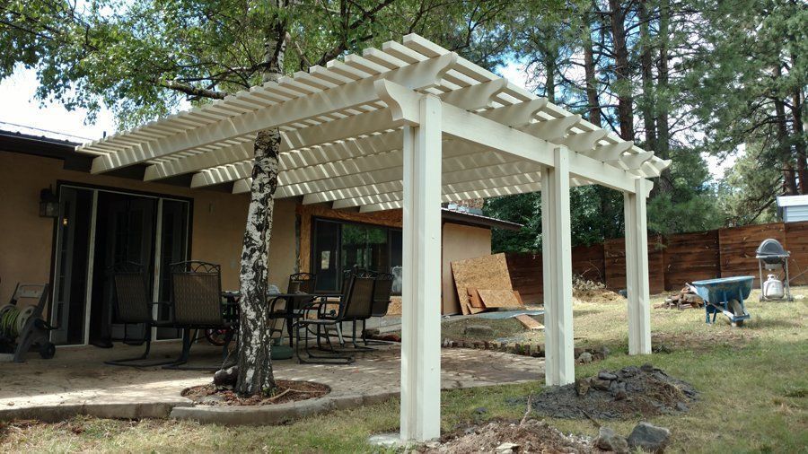 White pergola covers a patio, supported by columns. Trees and yard visible.