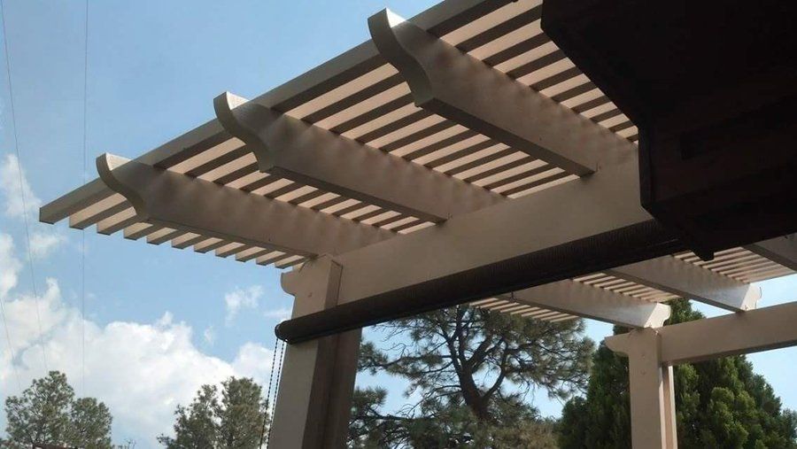 Pergola with white beams and slats against a blue sky, partially shaded by a roll-down screen.
