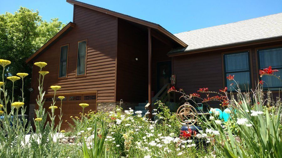 Brown house with a gable roof and flowers in the foreground on a sunny day.