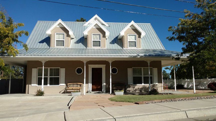 Tan house with a silver roof, three dormer windows, a porch, and a brown door on a sunny day.