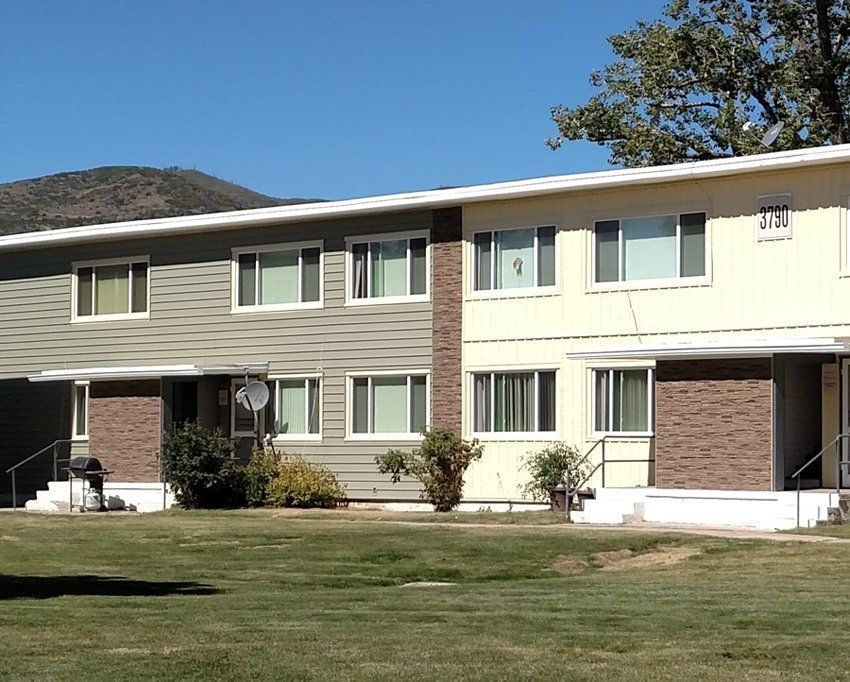 Two-story apartment building with green and beige siding. Brown brick accents, grass lawn, mountain in the background.