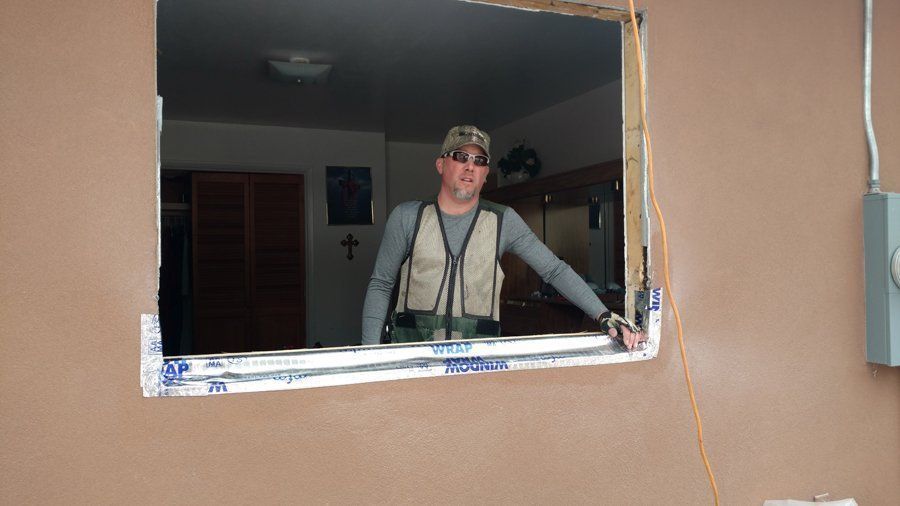 Man in work vest stands in a window frame. Brown wall, interior visible; room, art.