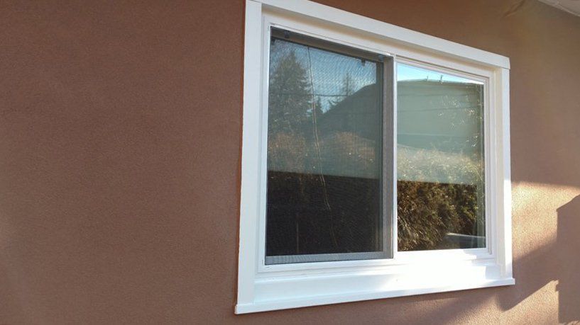 A sliding window with white trim on a brown stucco wall. The window reflects trees and sky.