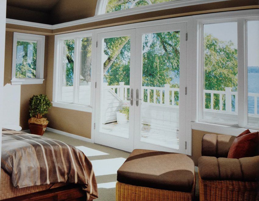 Bedroom with large white-framed windows overlooking a balcony and water, featuring furniture and sunlight.