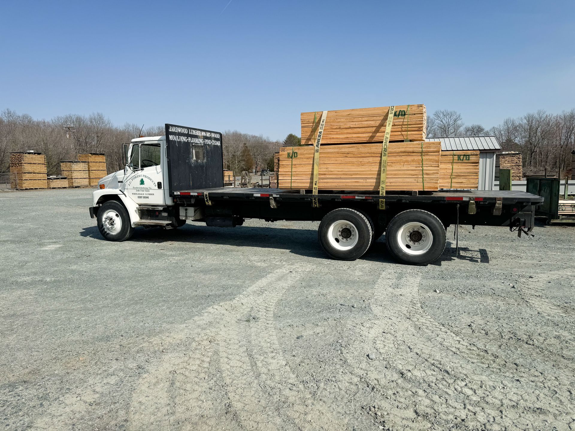 Flatbed truck carrying lumber on a gravel lot under a clear sky.