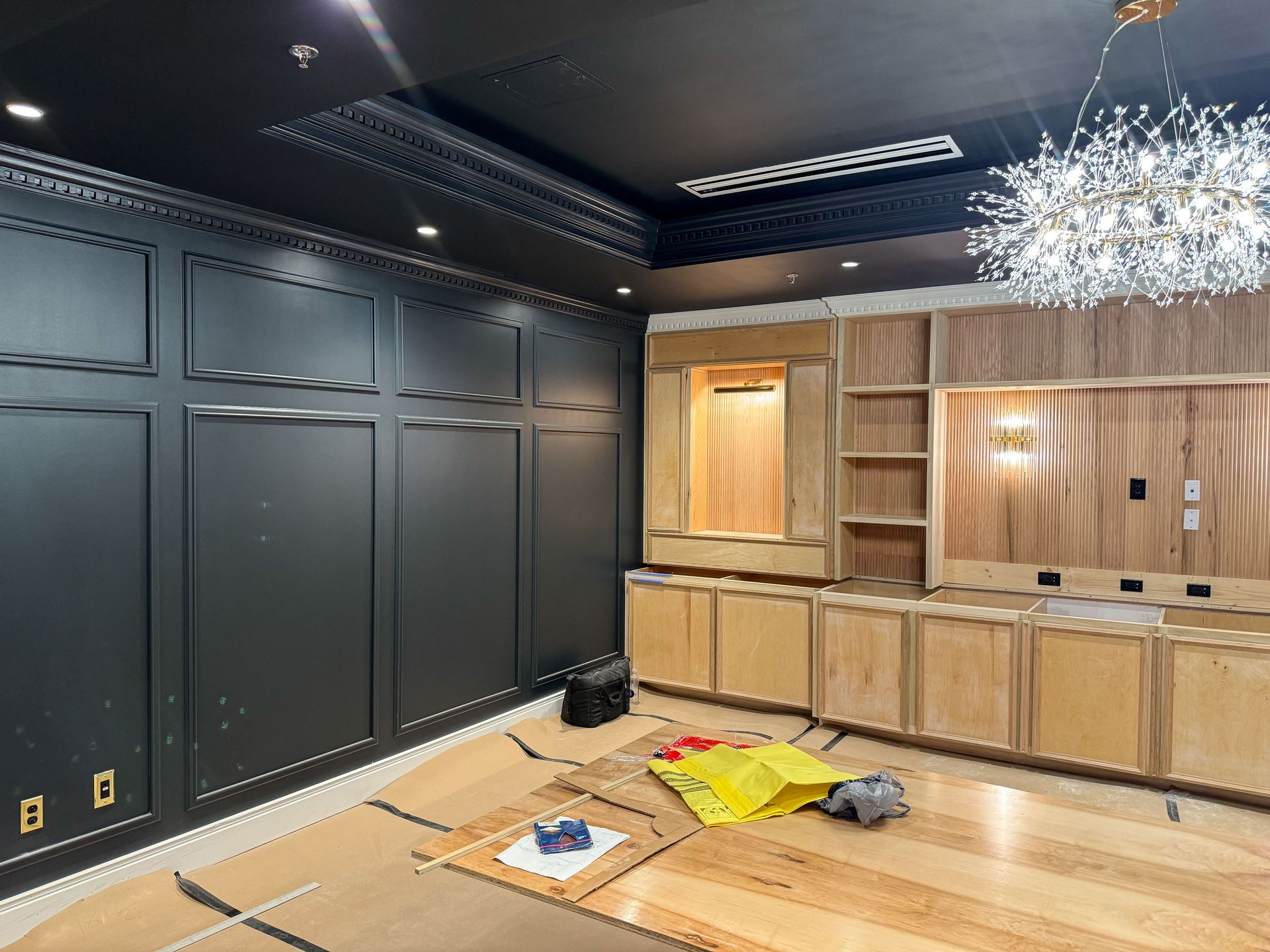 Room under construction with dark paneled walls, light wood cabinetry, and a chandelier.