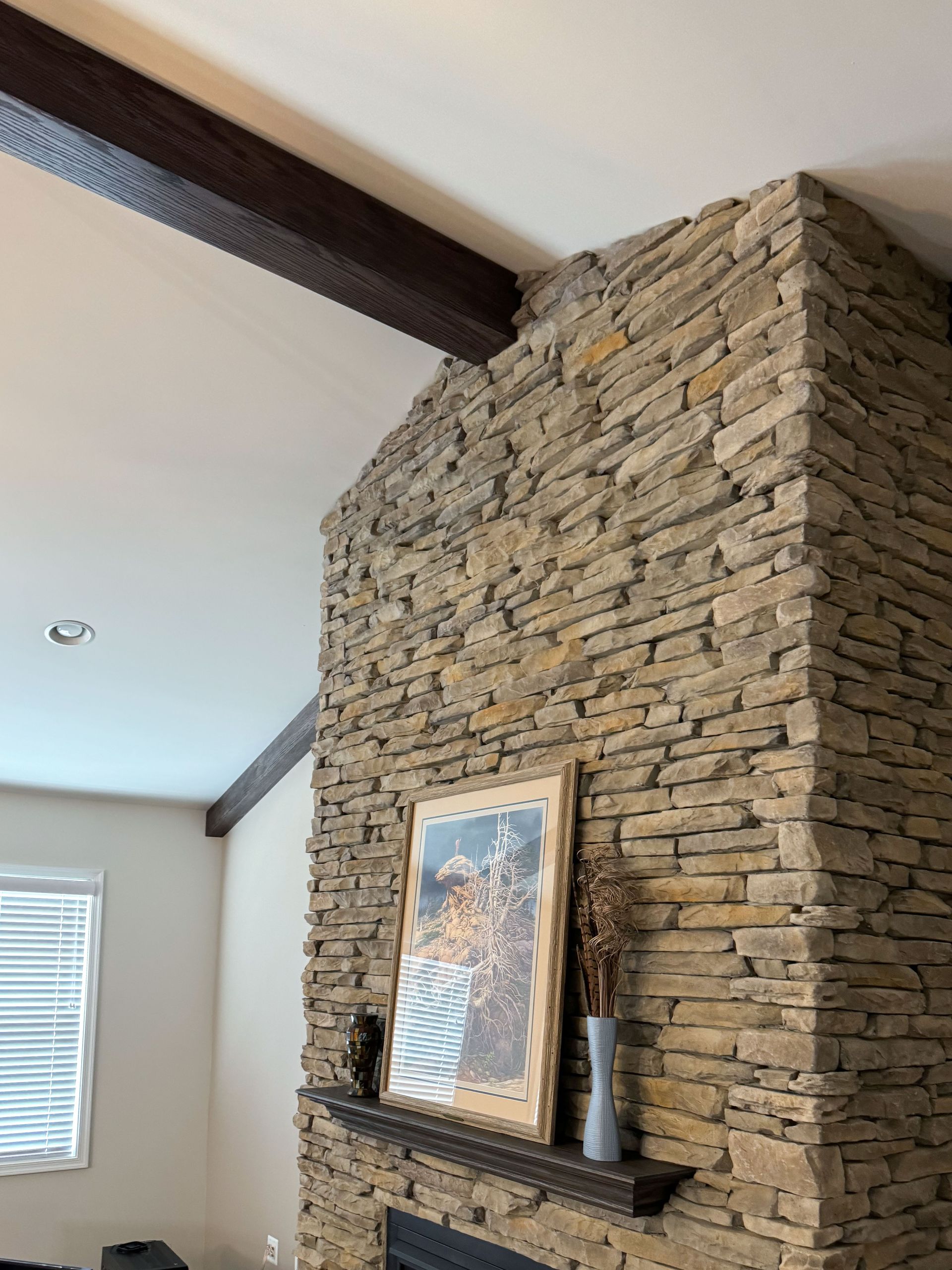 Stone fireplace with a wooden mantle and beam, artwork, and vase. Brown and beige tones, natural light.