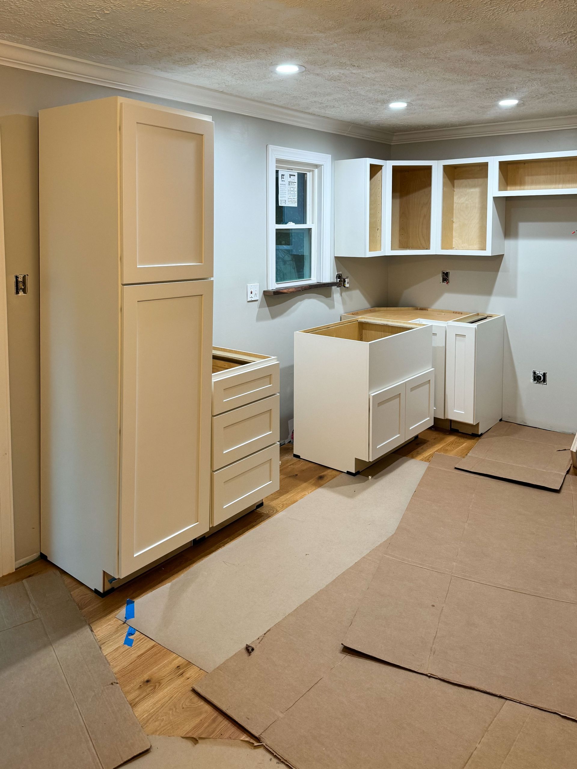 Kitchen under construction; white cabinets installed on light wood floor, beige walls, and drywall ceiling.