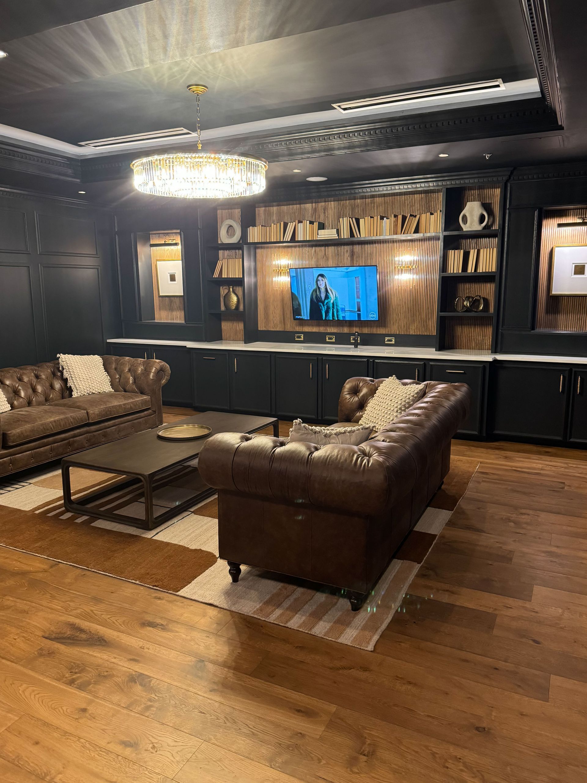 A luxurious den with leather sofas, a built-in entertainment center, and a chandelier; dark wood and black accents.