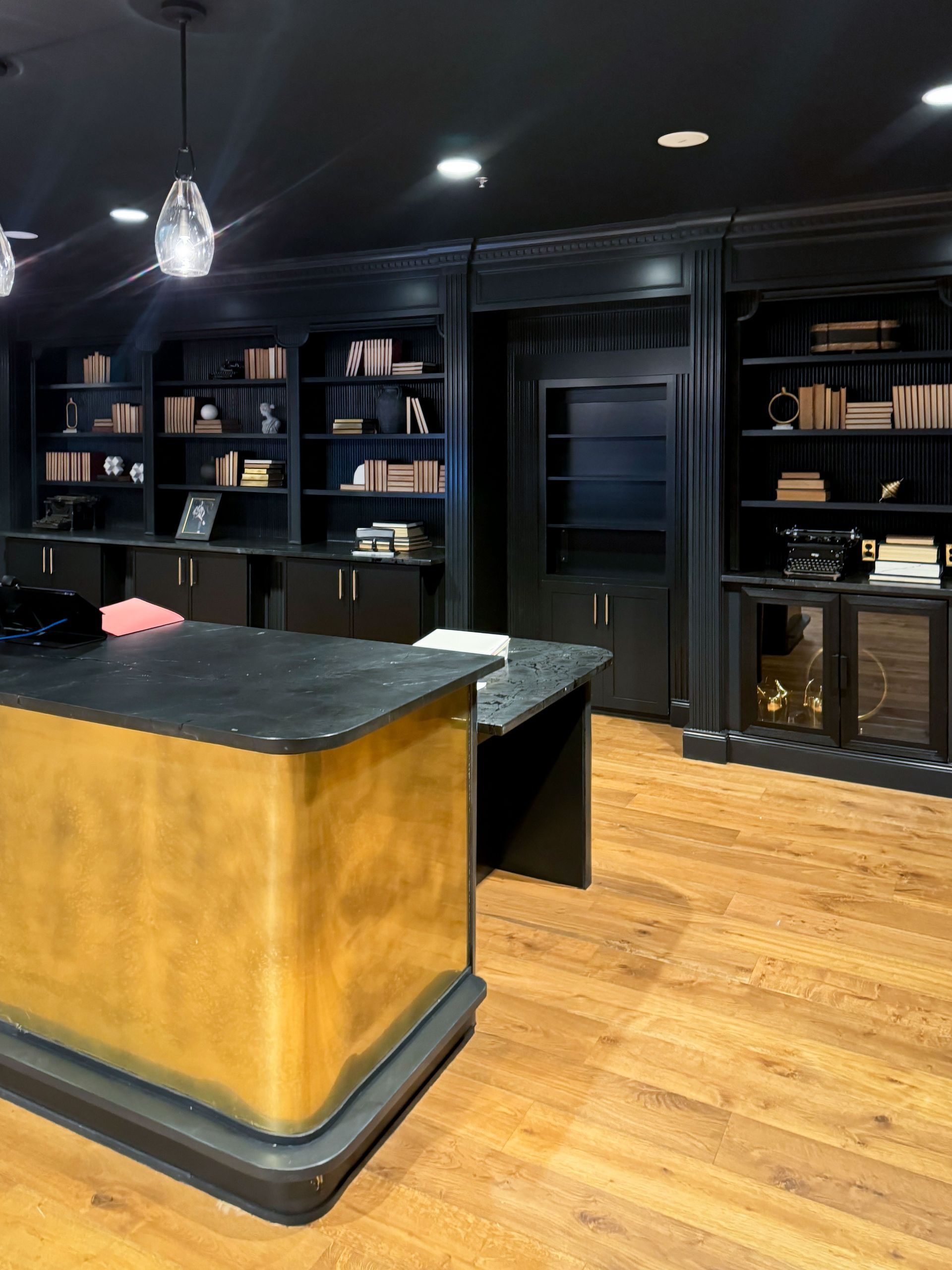 Dark room with black bookshelves, golden desk, wood floor, and hanging lights.