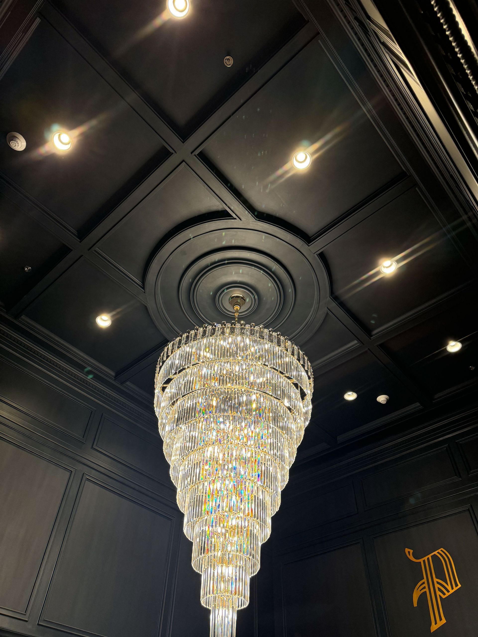 Ornate crystal chandelier hangs from a dark, decorative ceiling with recessed lighting.