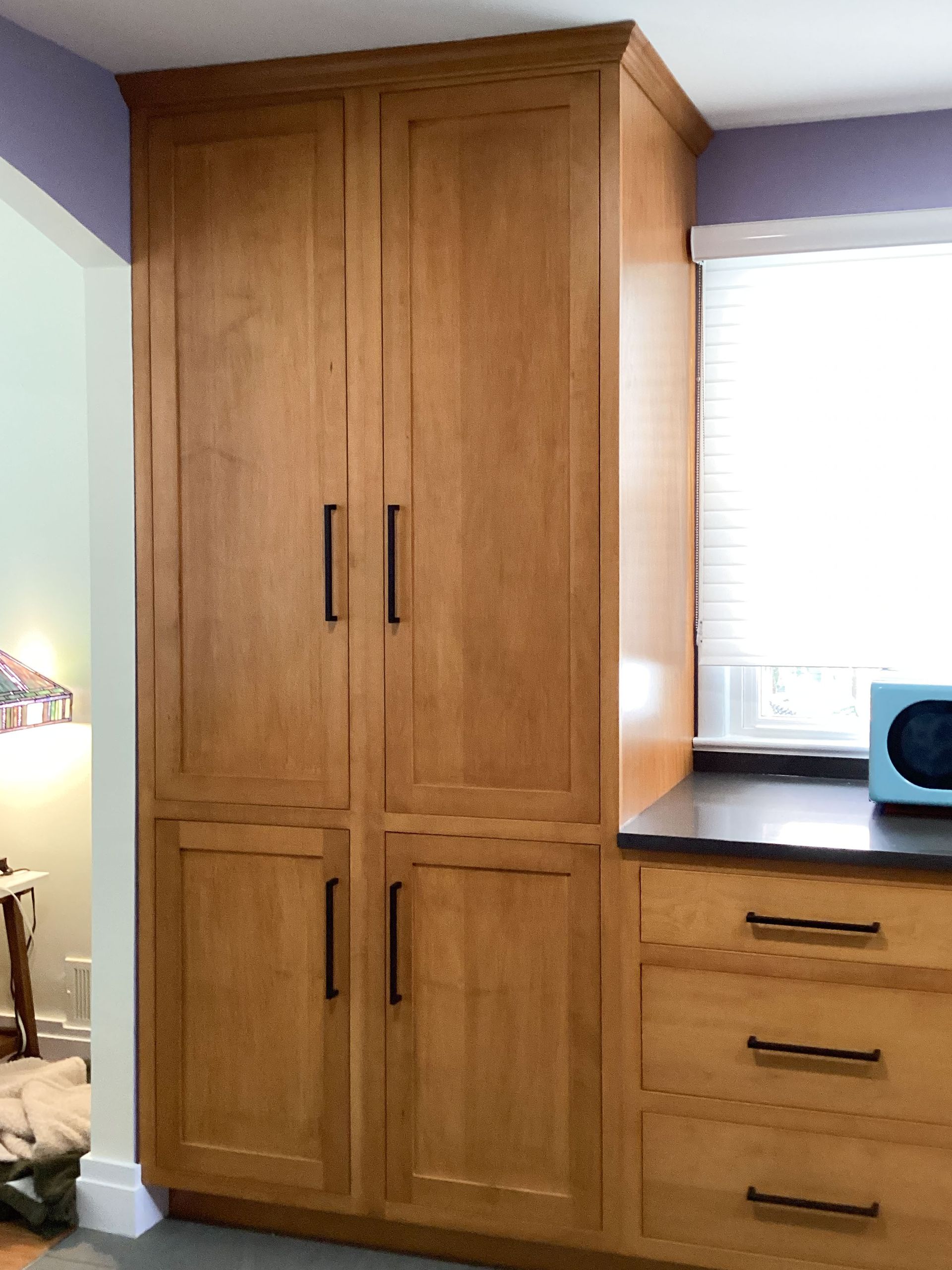 Tall wooden pantry cabinet with black handles, next to a countertop and window.
