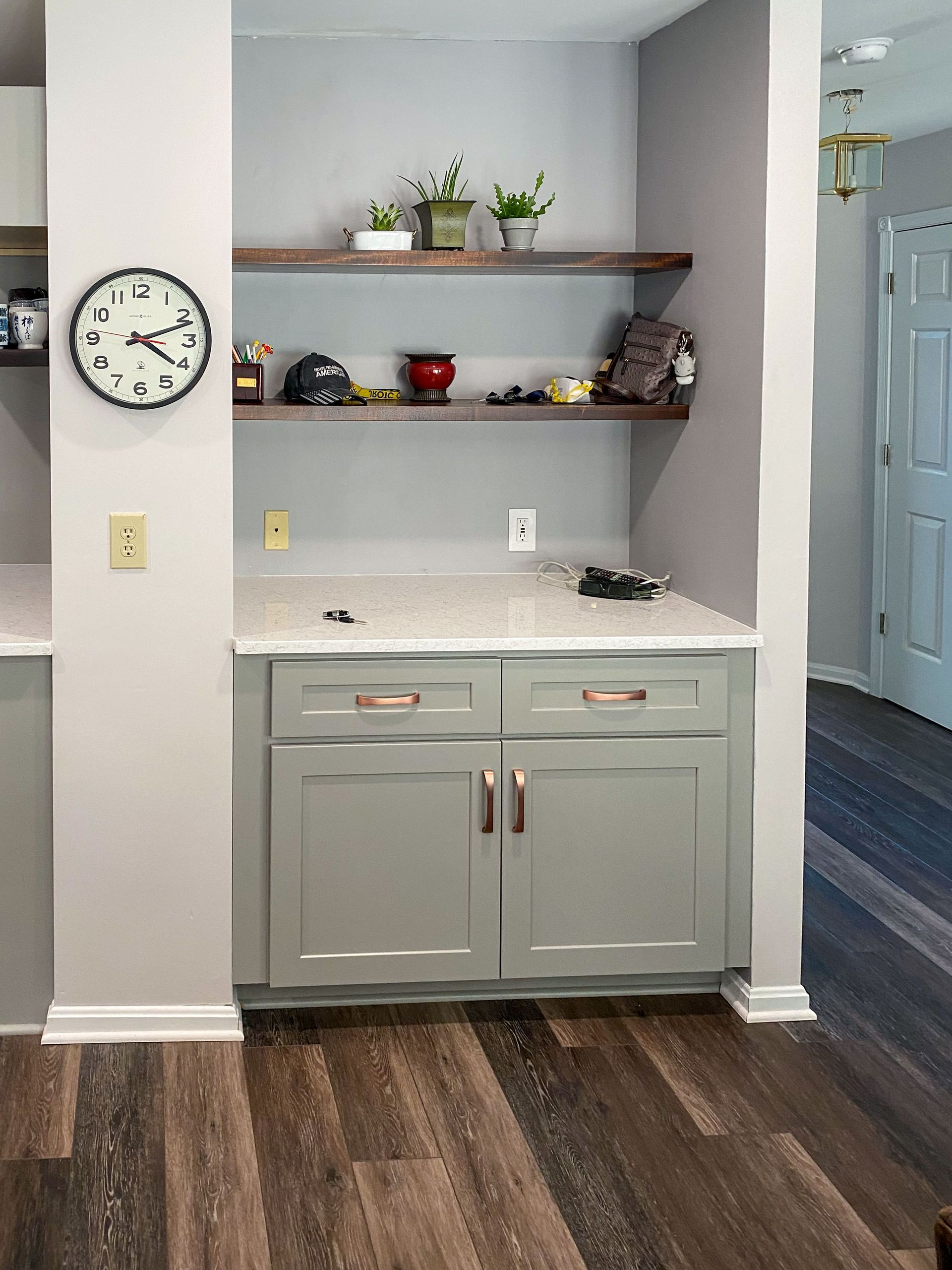 Built-in kitchen storage: gray cabinets, stone countertop, two wooden shelves with decorative items, clock on wall.