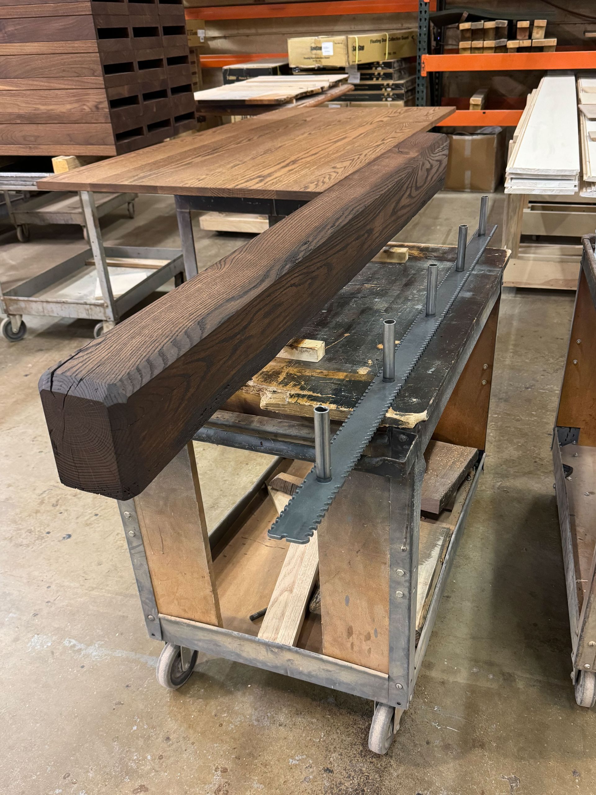 A dark wooden beam rests on a cart with metal supports. Workshop setting with other lumber in the background.