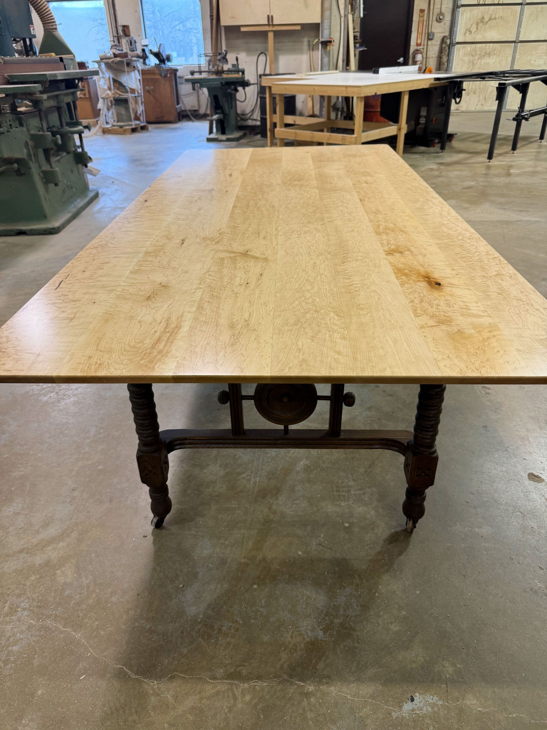 Wooden table with a light-colored top and a dark, ornate base with wheels, in a workshop.