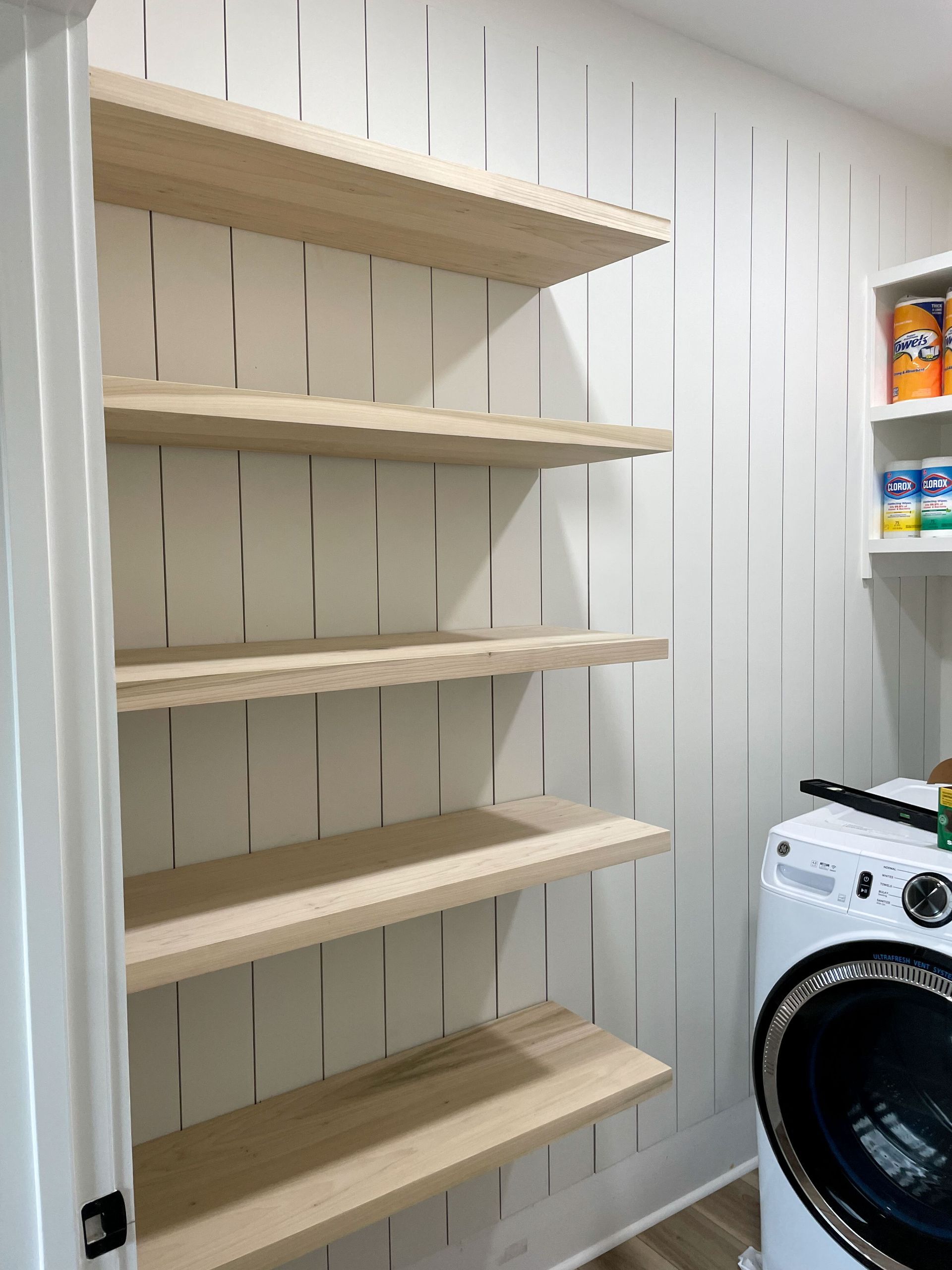 Laundry room with vertical-striped wall and wooden shelves. White walls, a washing machine, and cleaning supplies.