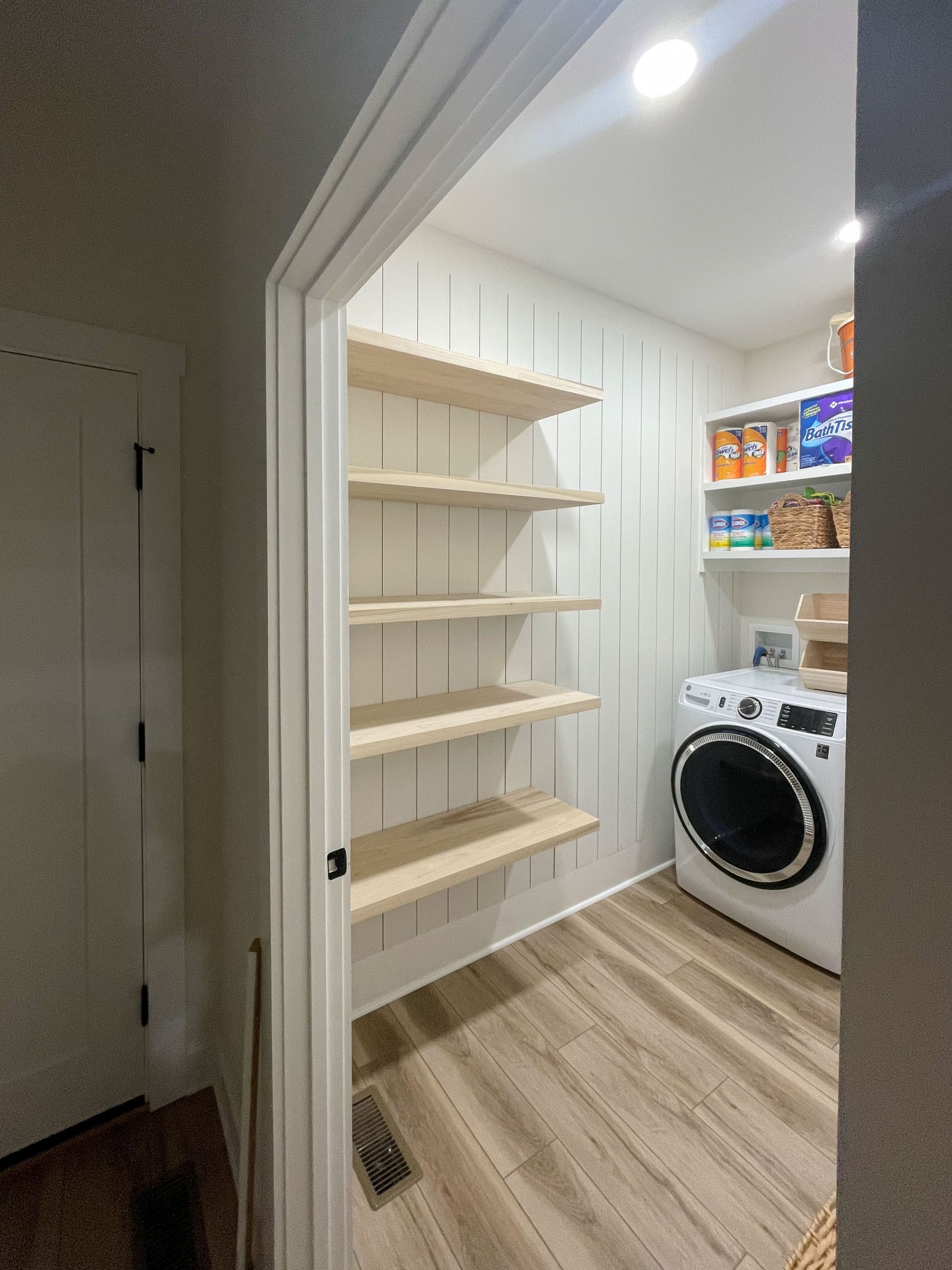 Pantry with wooden shelves, shiplap walls, and a washing machine. Doorway visible.