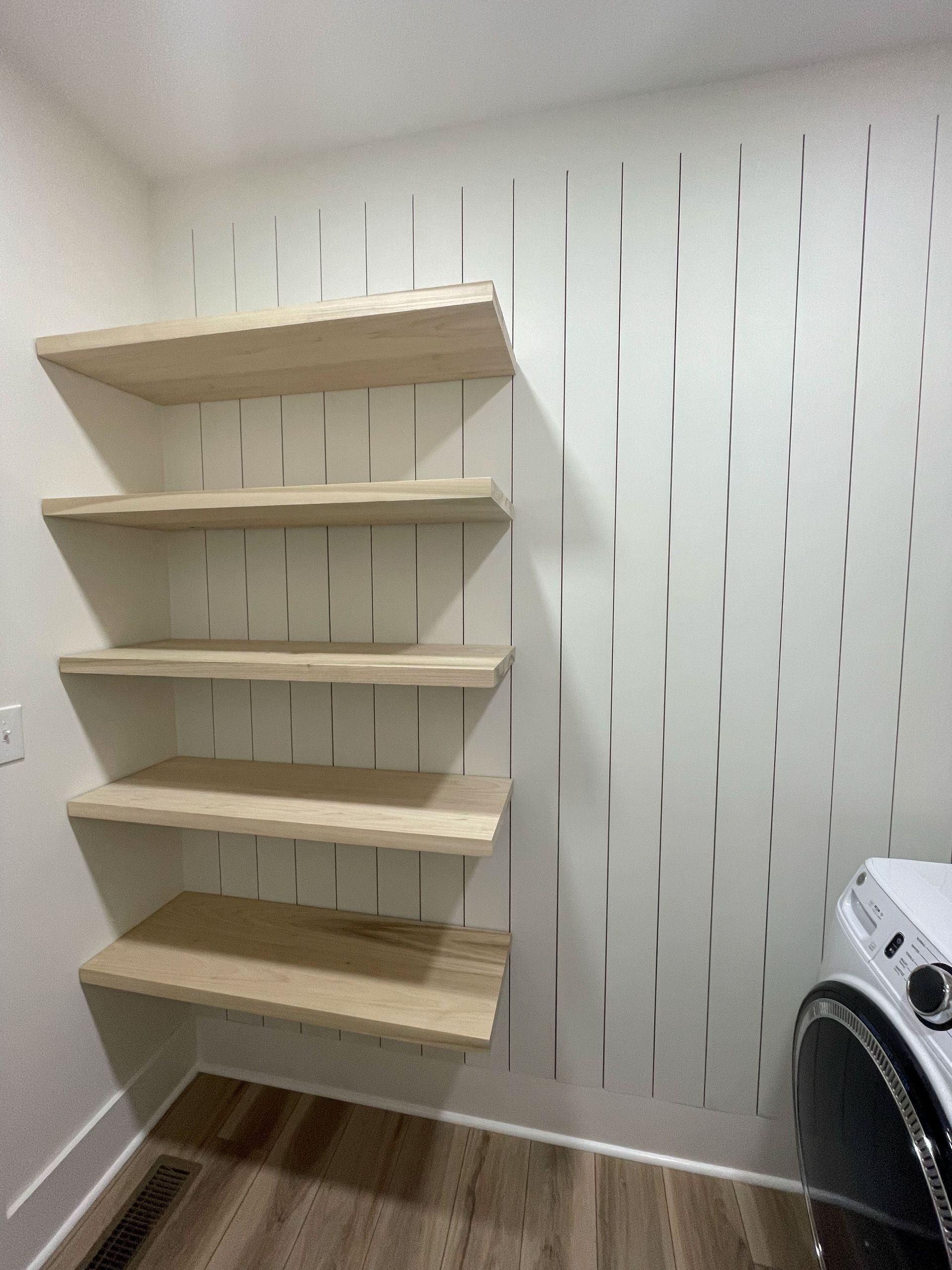 Floating wooden shelves on a white wall with a decorative, vertical design. A washing machine sits nearby.