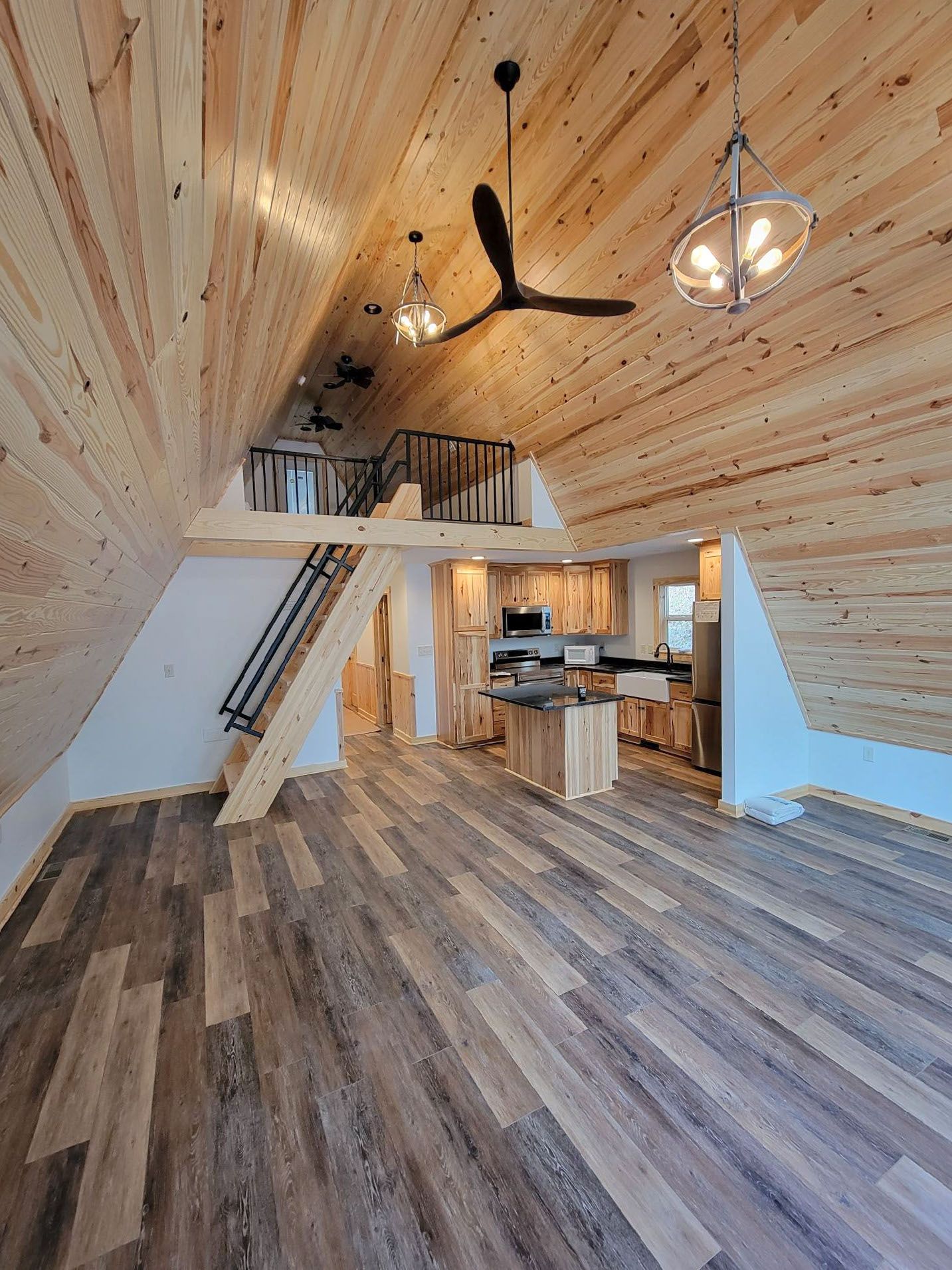 Interior of a cabin-like home with wood-paneled walls and a loft. Kitchen and living area with a dark ceiling fan and hanging lights.