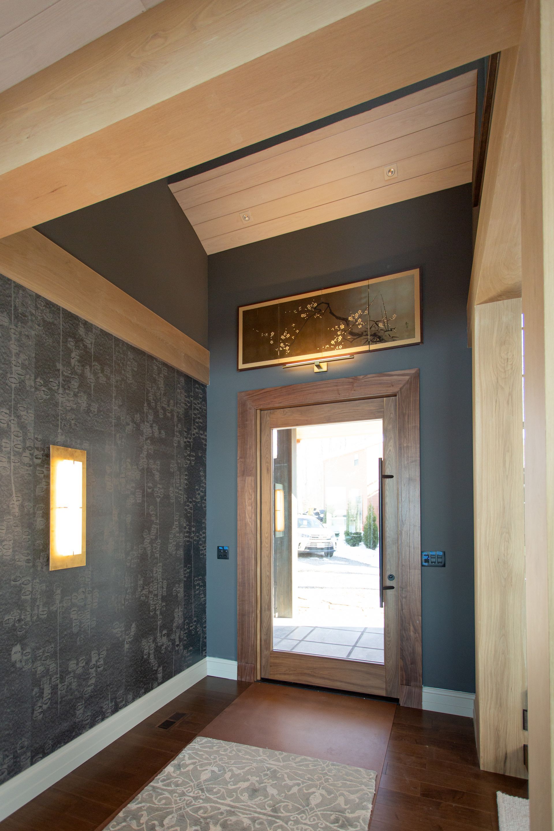 Entryway with gray textured wall, wooden door, and gold-framed art. Light wood beams and trim frame the space.