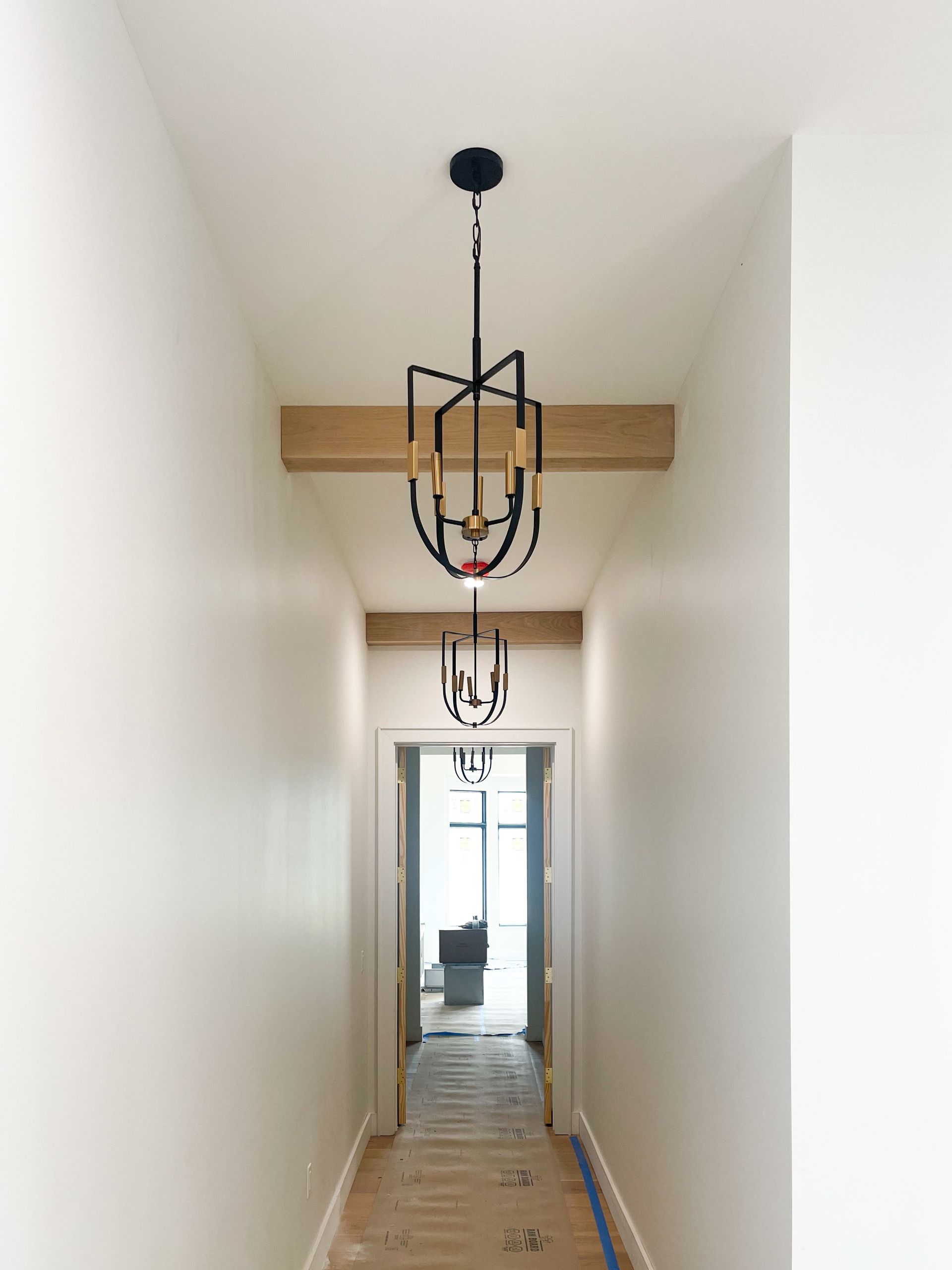 Narrow hallway with two black chandeliers, wooden beams, and white walls.